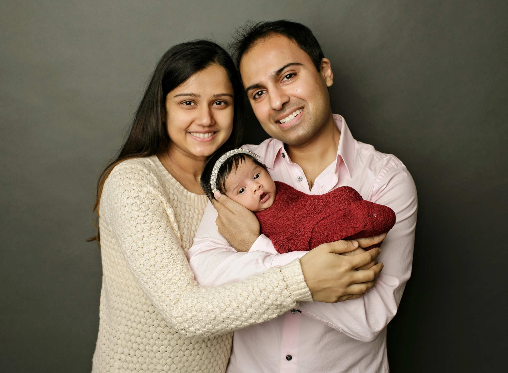 Two parents smiling while holding their infant wrapped in a red blanket