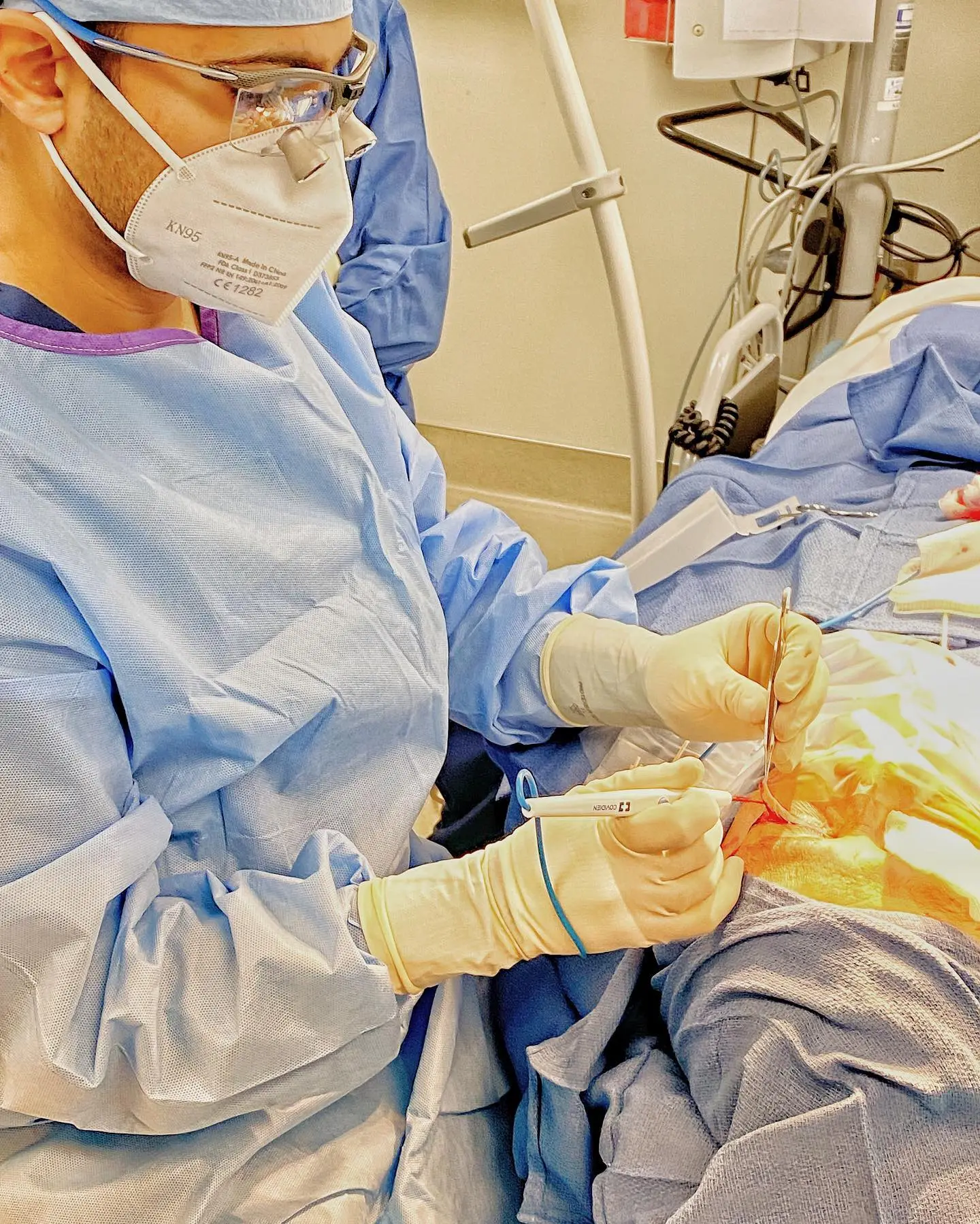 Surgeon in blue gown and yellow gloves performing eyelid surgery on patient under bright operating lights