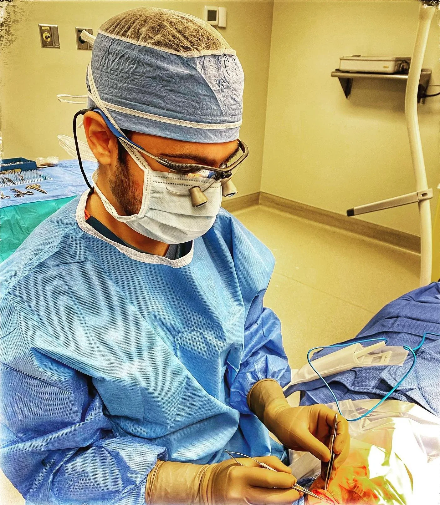 Surgeon in blue gown, headwrap, mask, and glasses performing procedure in yellow-walled operating room
