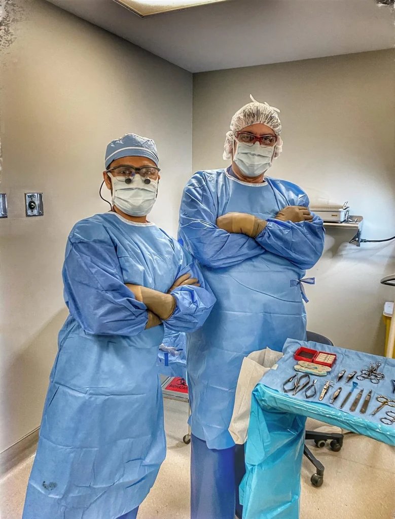 Two surgeons in blue surgical gowns, masks, and head coverings standing with arms crossed in operating room with surgical instruments on table