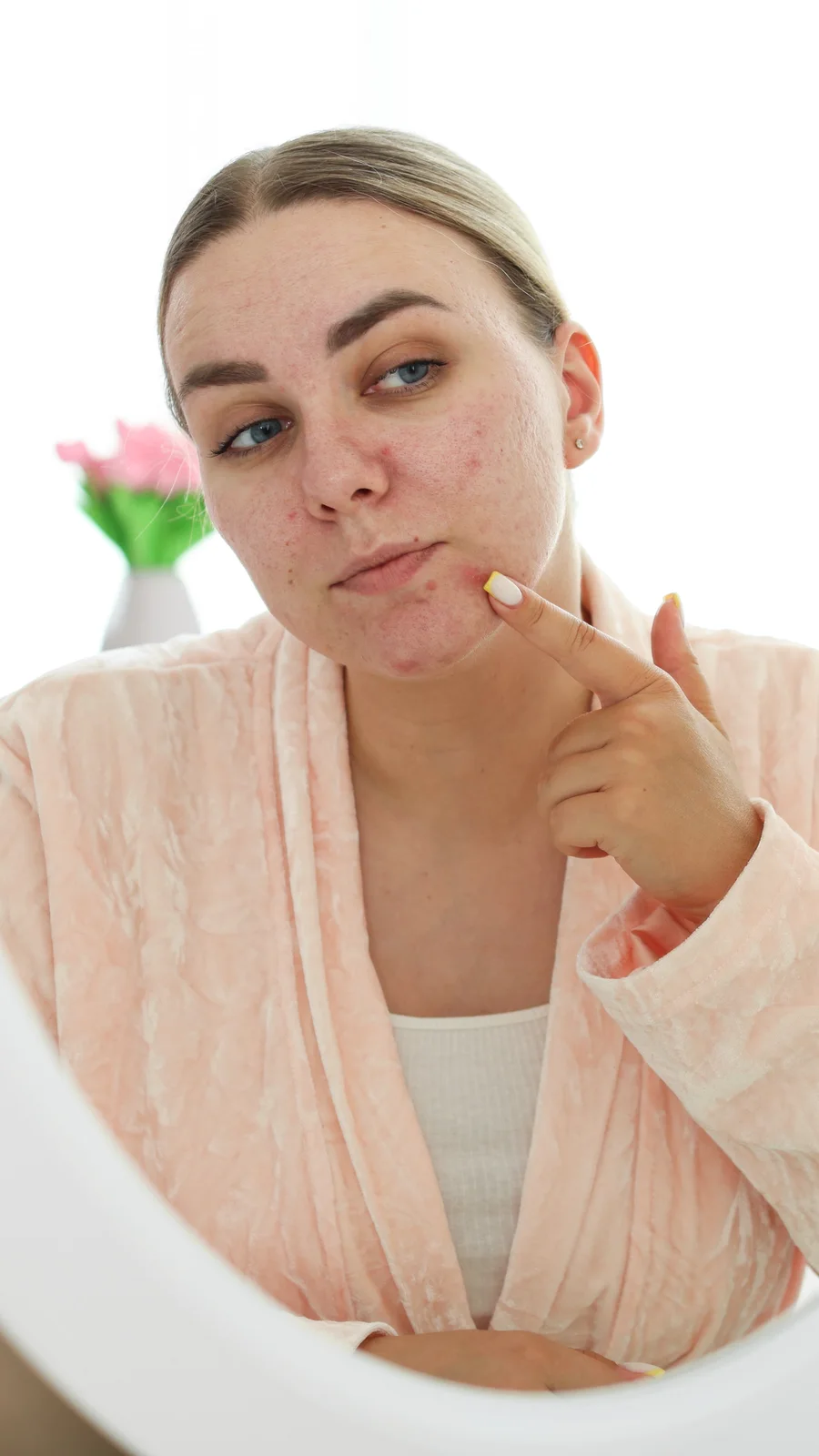Woman in a soft pink robe checking small white bumps on her face in a vanity mirror
