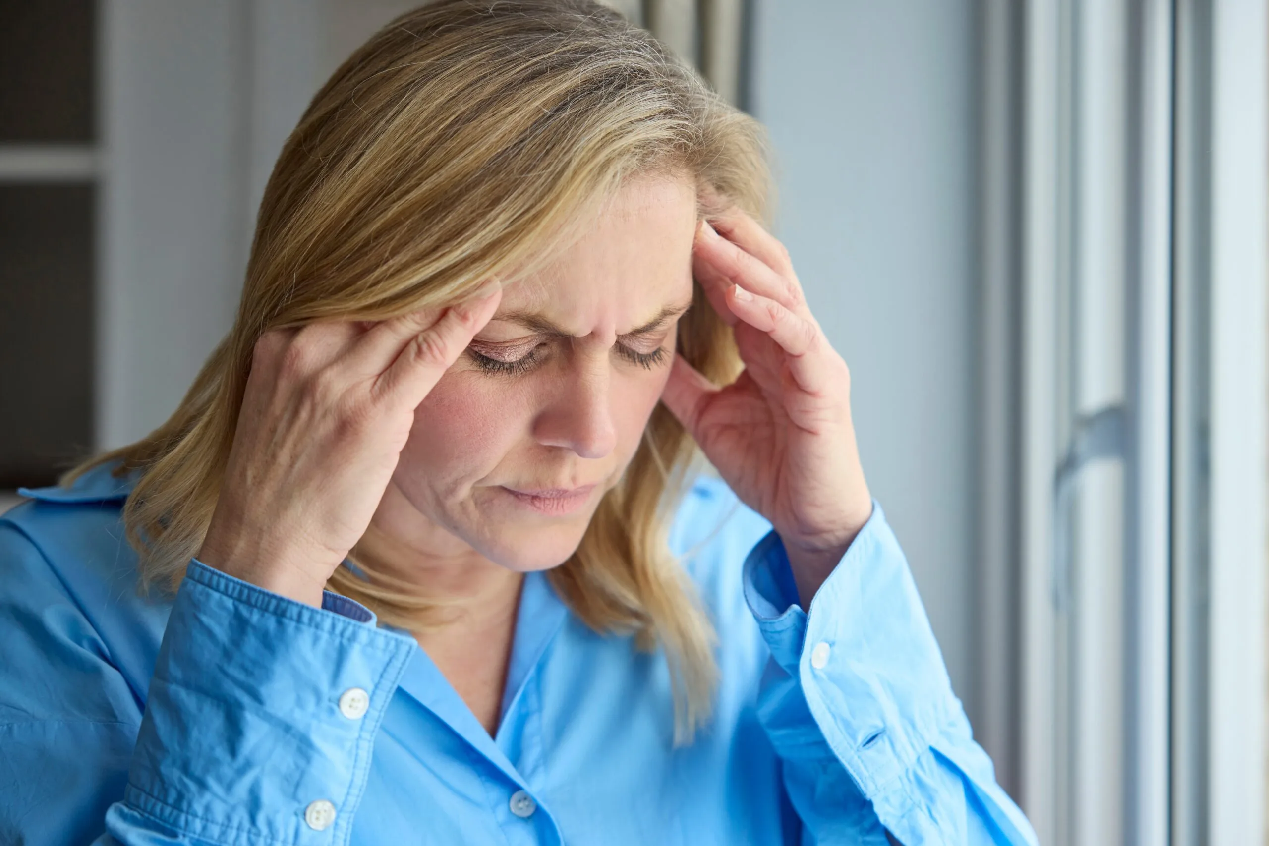Woman in blue shirt holding head with both hands experiencing headache