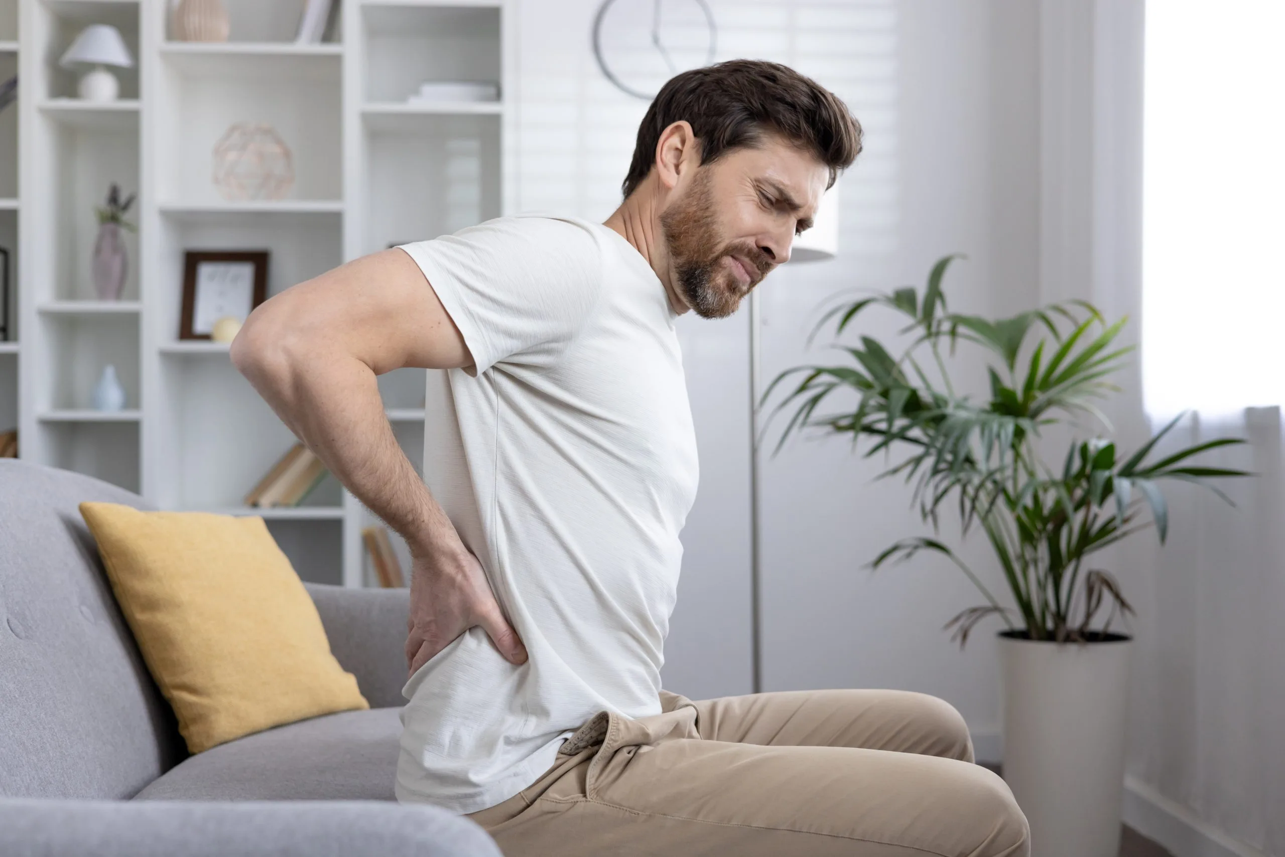 Man in white shirt holding lower back while sitting on couch in home living room