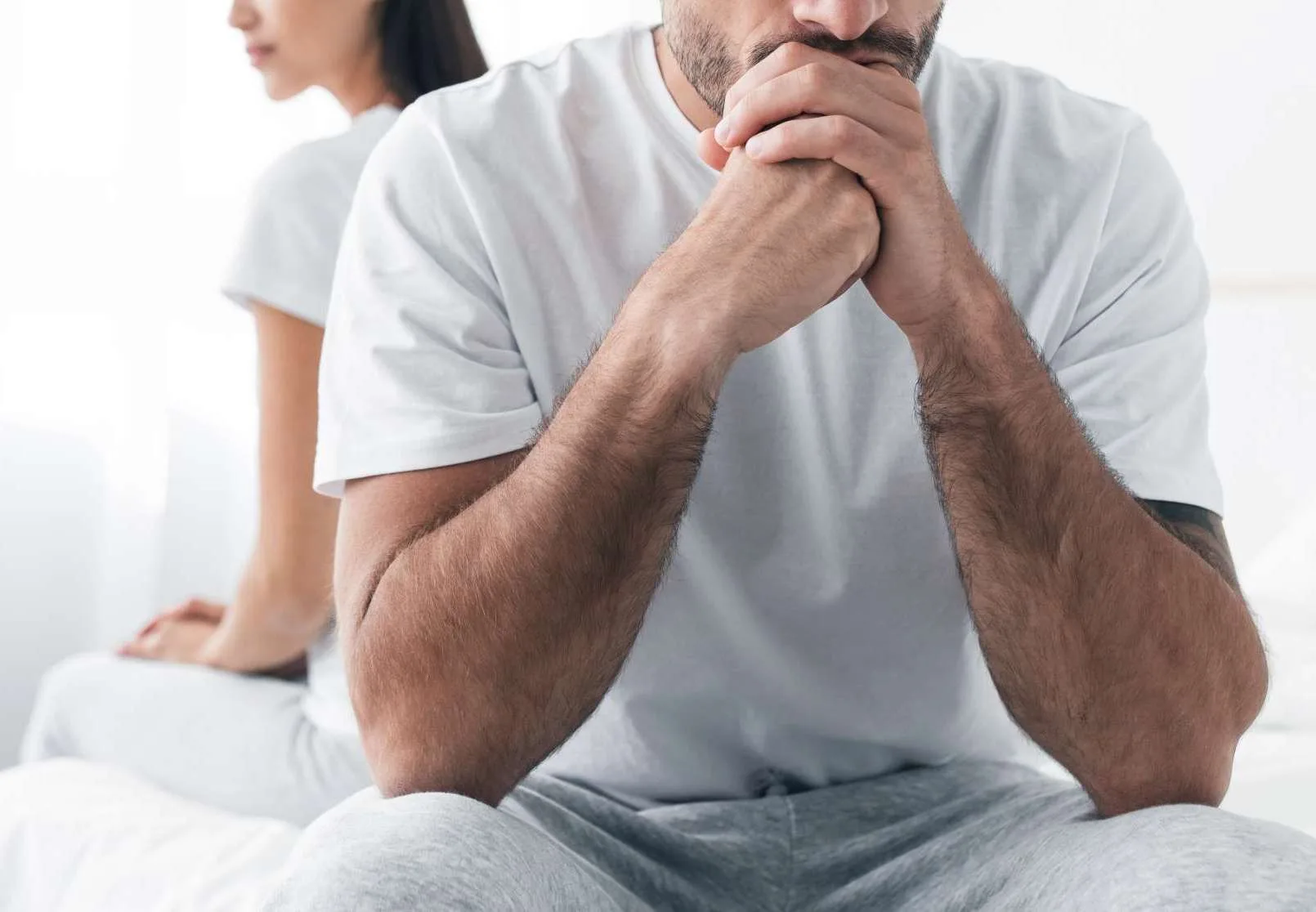 Man sitting on bed with hand to face, looking distressed, while woman sits beside him in background