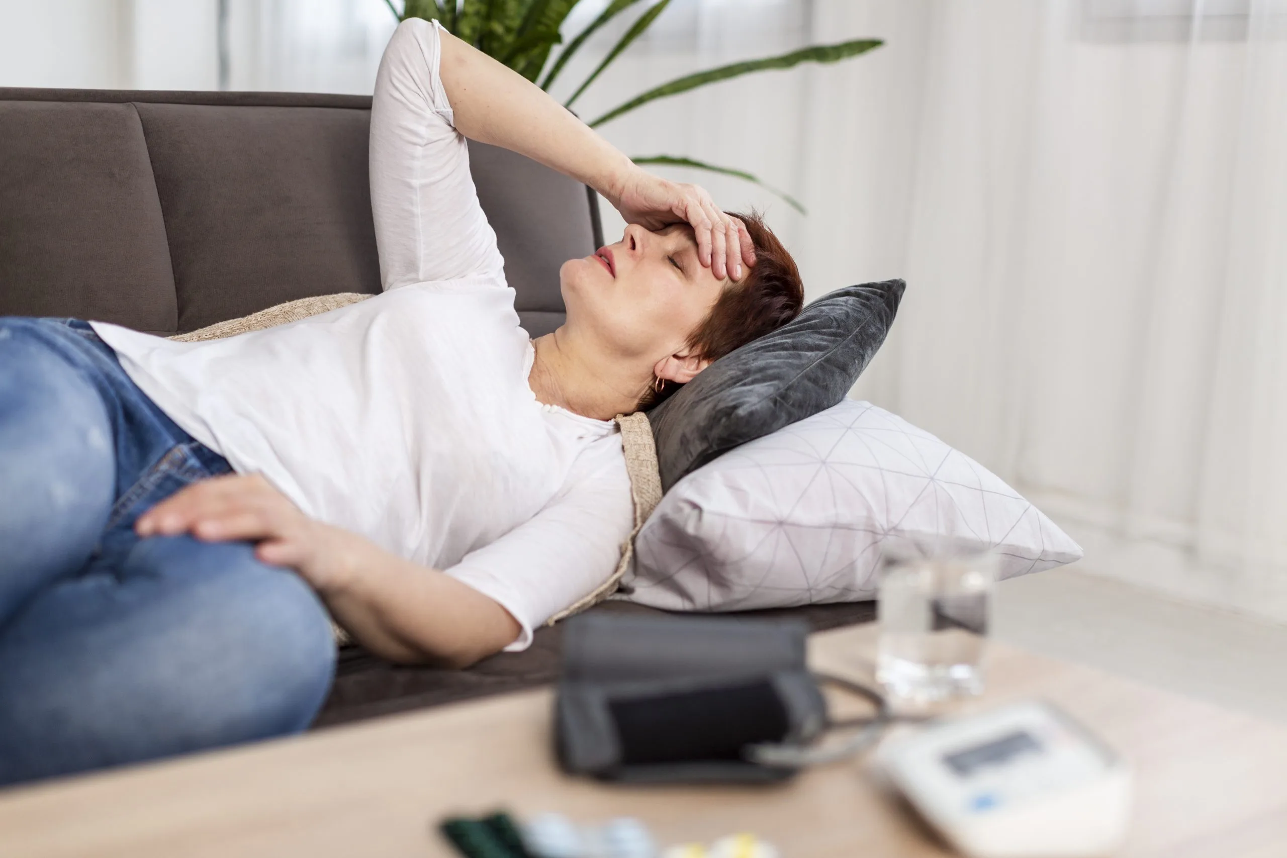 Senior woman resting on couch with hand to head, blood pressure monitor and medications on table nearby