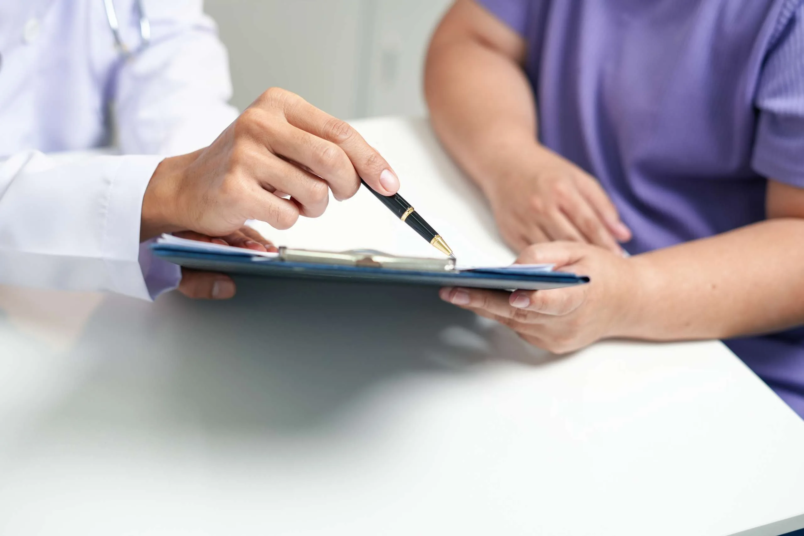 Doctor in white coat pointing to clipboard while consulting with patient in purple shirt