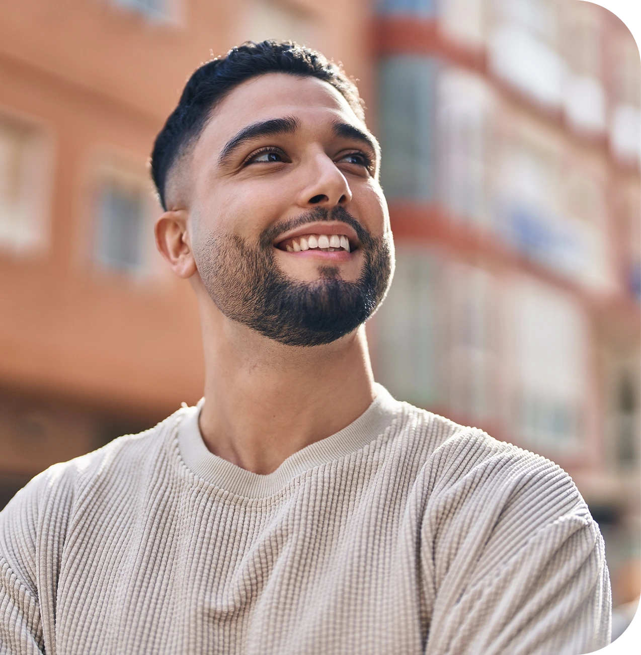 Smiling man wearing a light beige ribbed shirt outdoors with brick buildings blurred in background