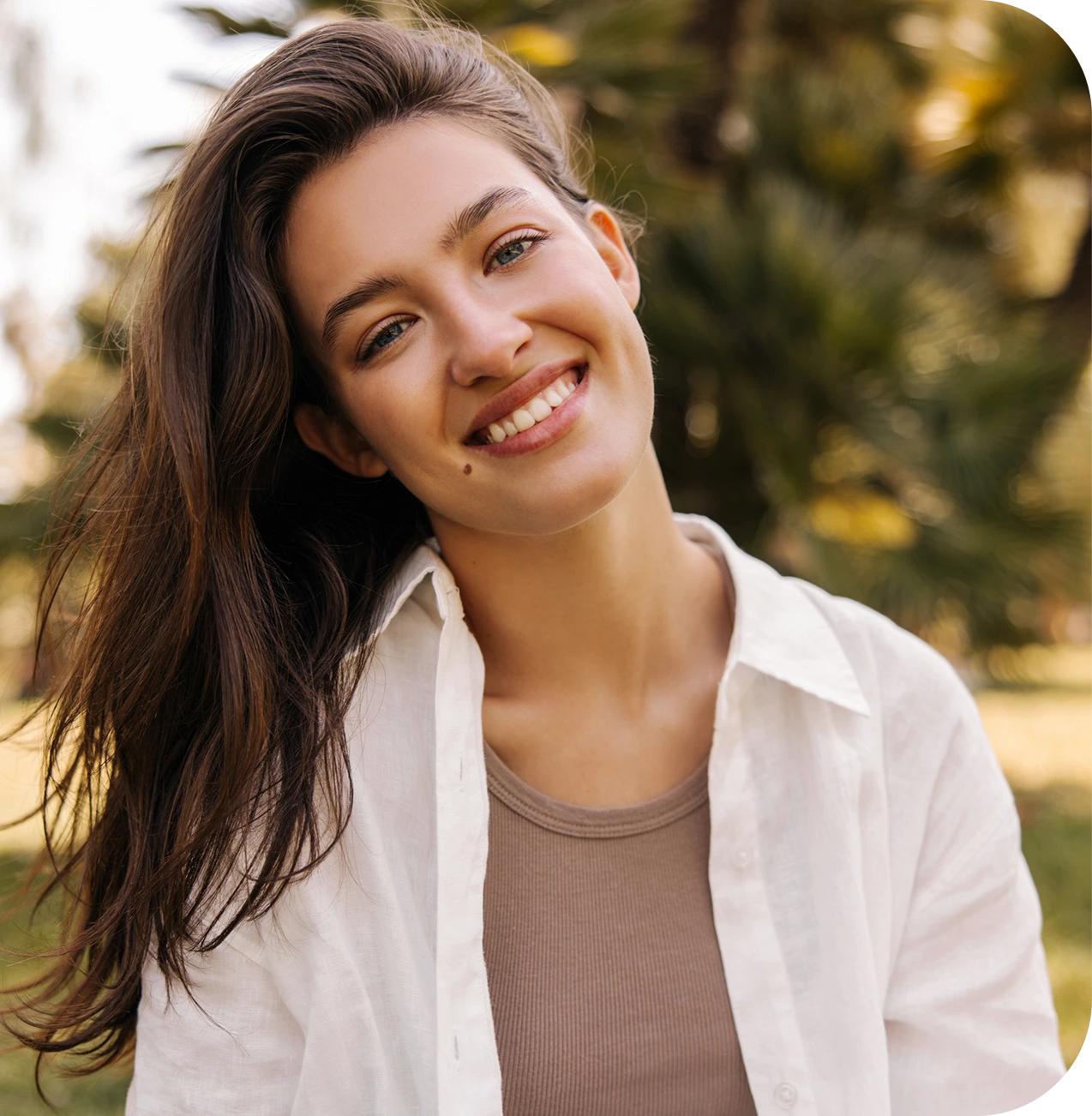 Woman smiling outdoors wearing white cardigan over gray top with blurred green foliage background