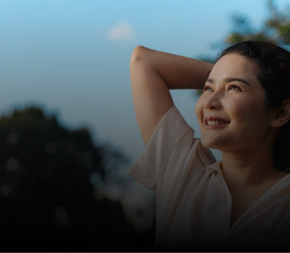 Woman smiling outdoors with hand near head, looking upward in peaceful expression