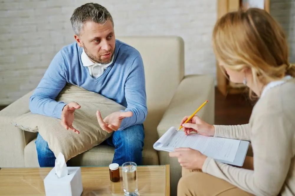 Adult man in blue sweater discussing ADHD treatment with a therapist holding a clipboard during a consultation