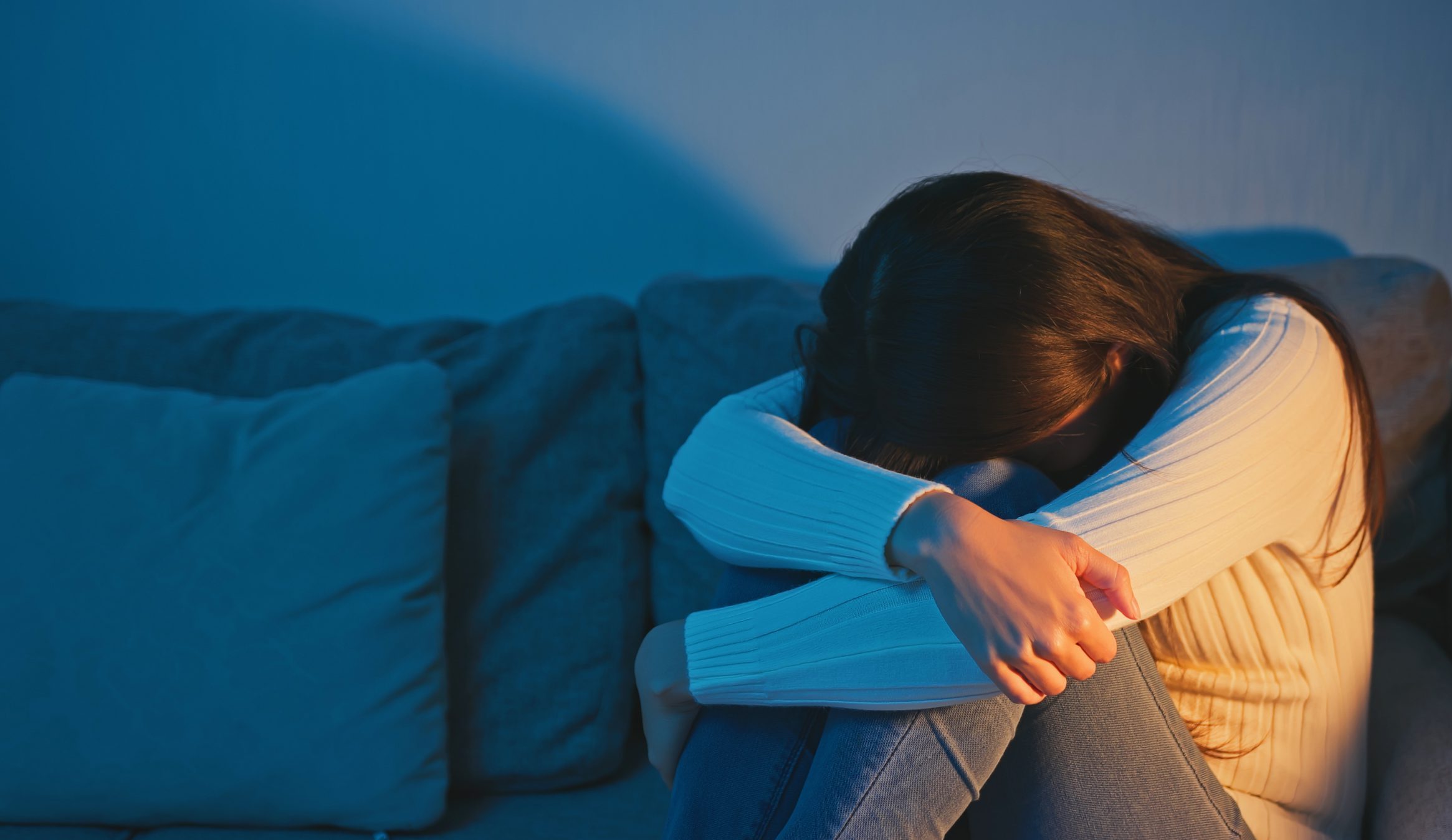 Person sitting on bed hugging knees in dim blue light, showing emotional distress