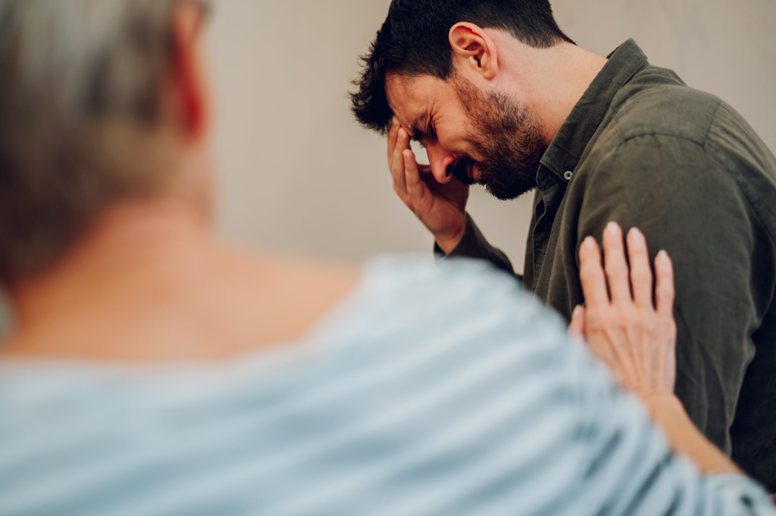 Man in counseling session holding his face while receiving supportive hand on shoulder from therapist