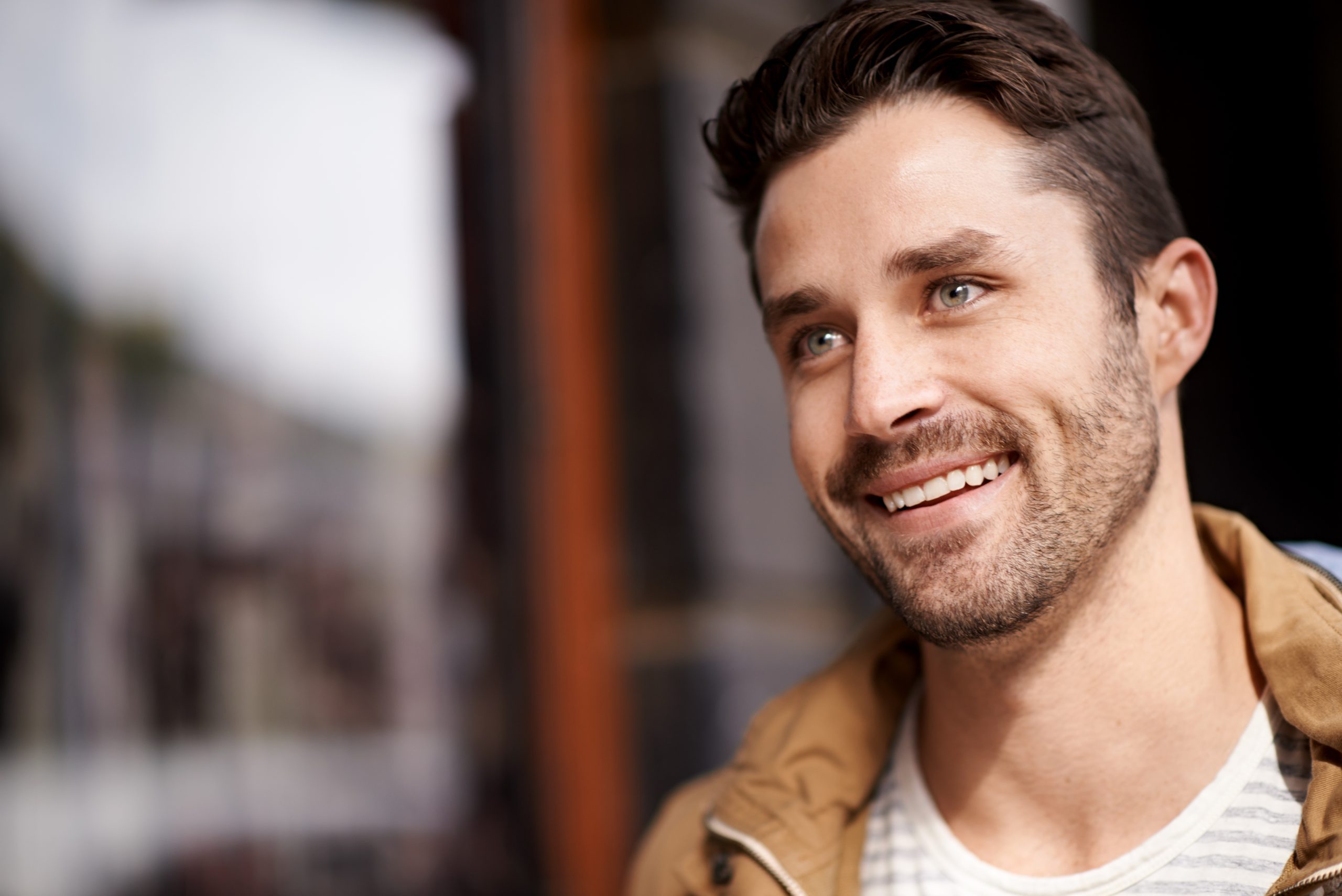 Man in tan jacket smiling at camera outdoors with blurred background