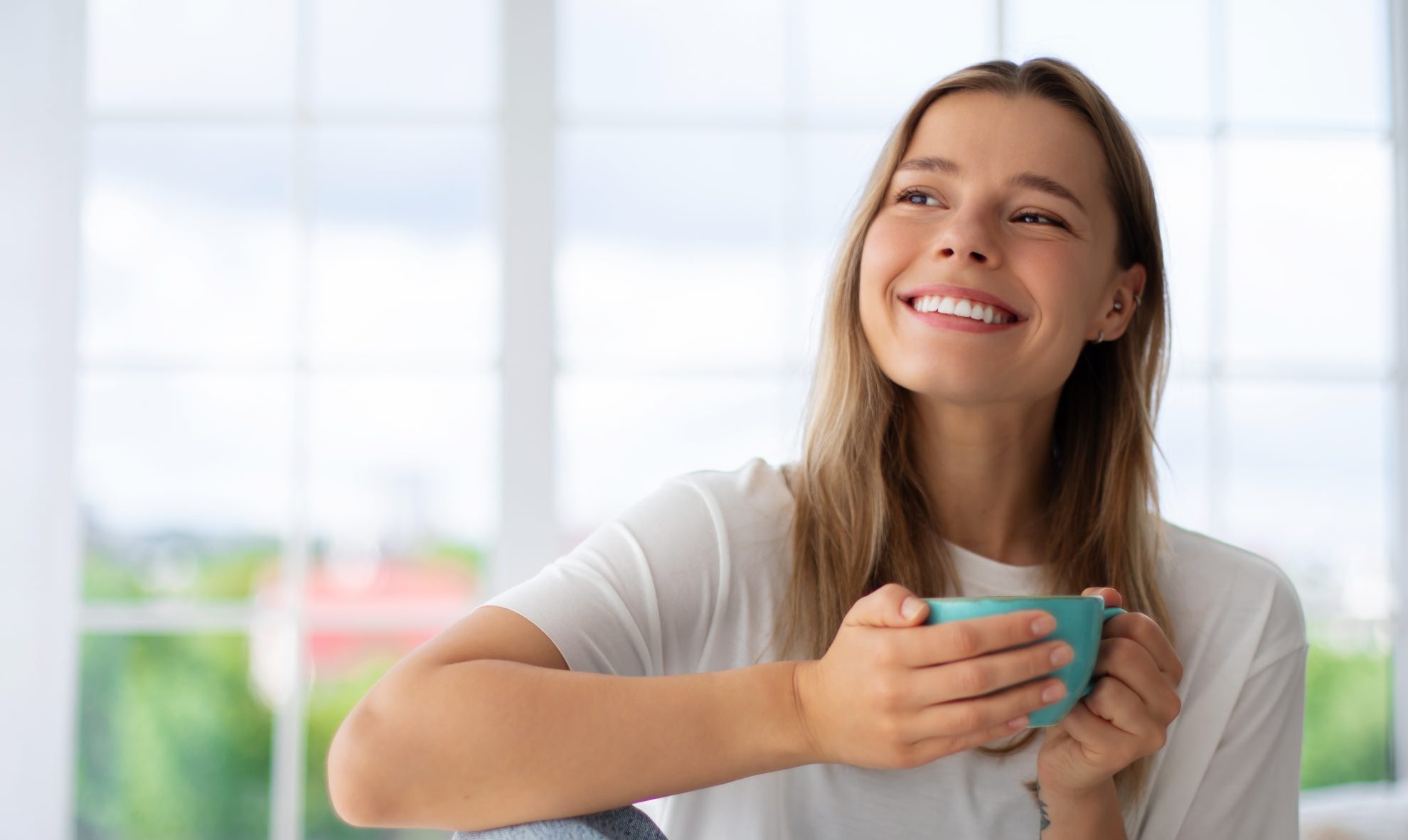 Woman holding teal mug smiling while looking out bright window with city view