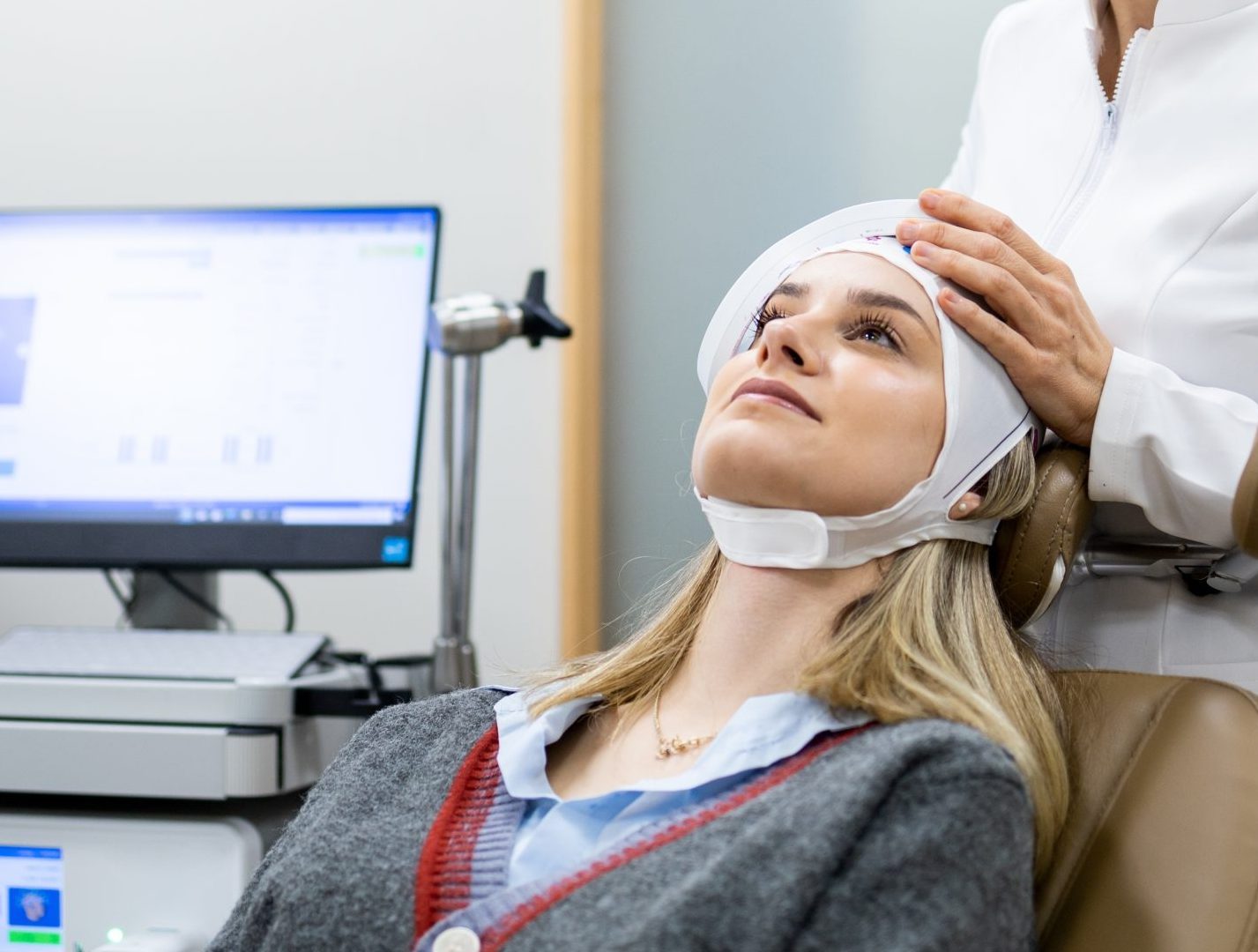 Clinician adjusting white head wrap on patient during facial treatment with computer monitor visible