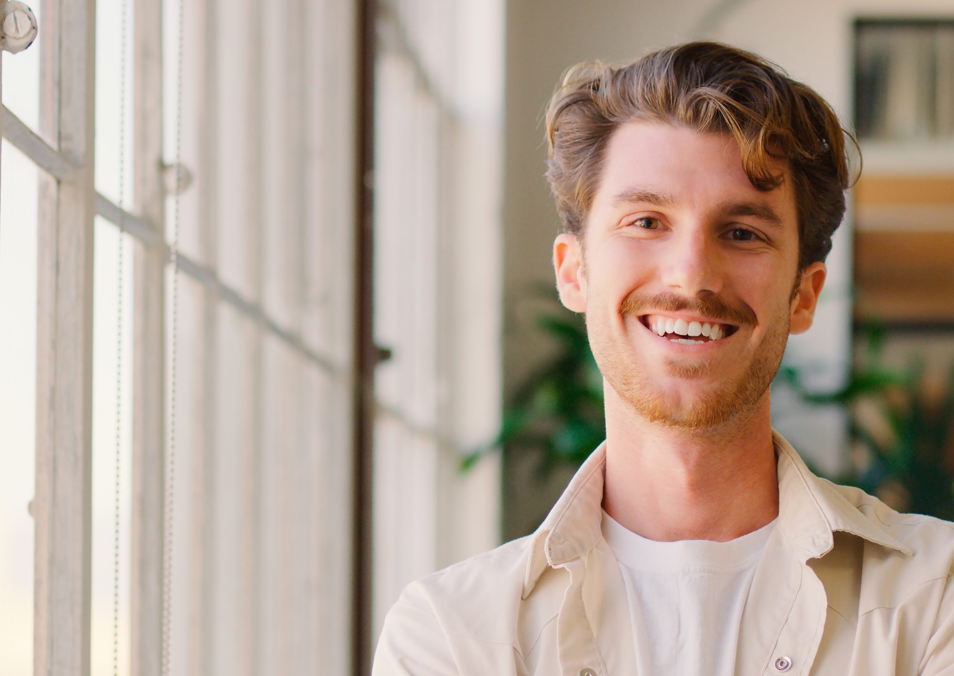 Man in beige shirt smiling by bright window with plants visible