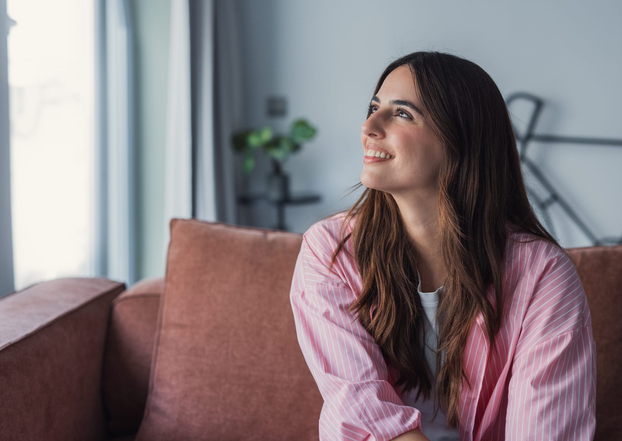 Woman with dark hair wearing pink striped shirt, sitting on couch looking toward window