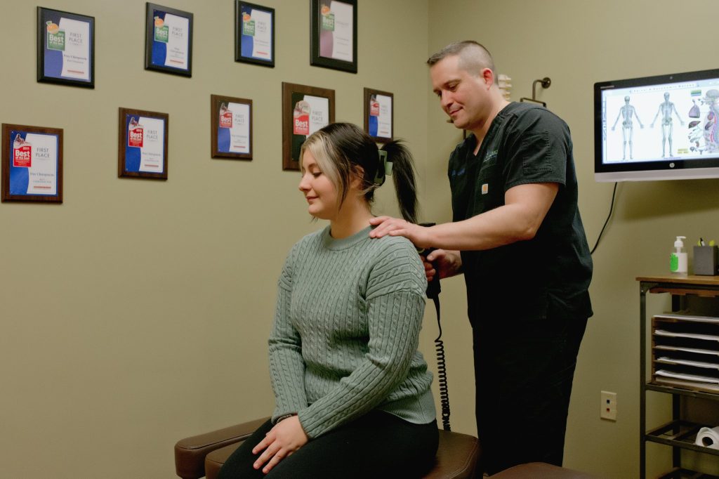 Chiropractor examining patient's shoulder in treatment room with anatomical charts on wall