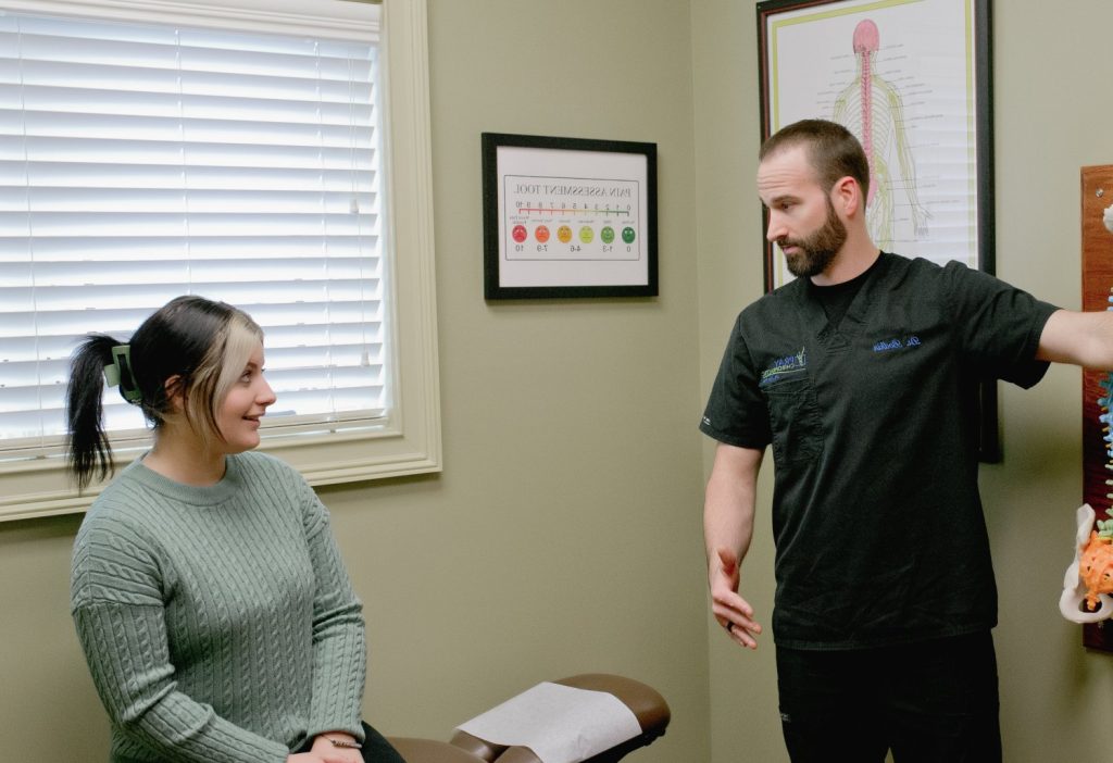 Chiropractor in dark shirt discussing spinal anatomy chart with seated patient in treatment room