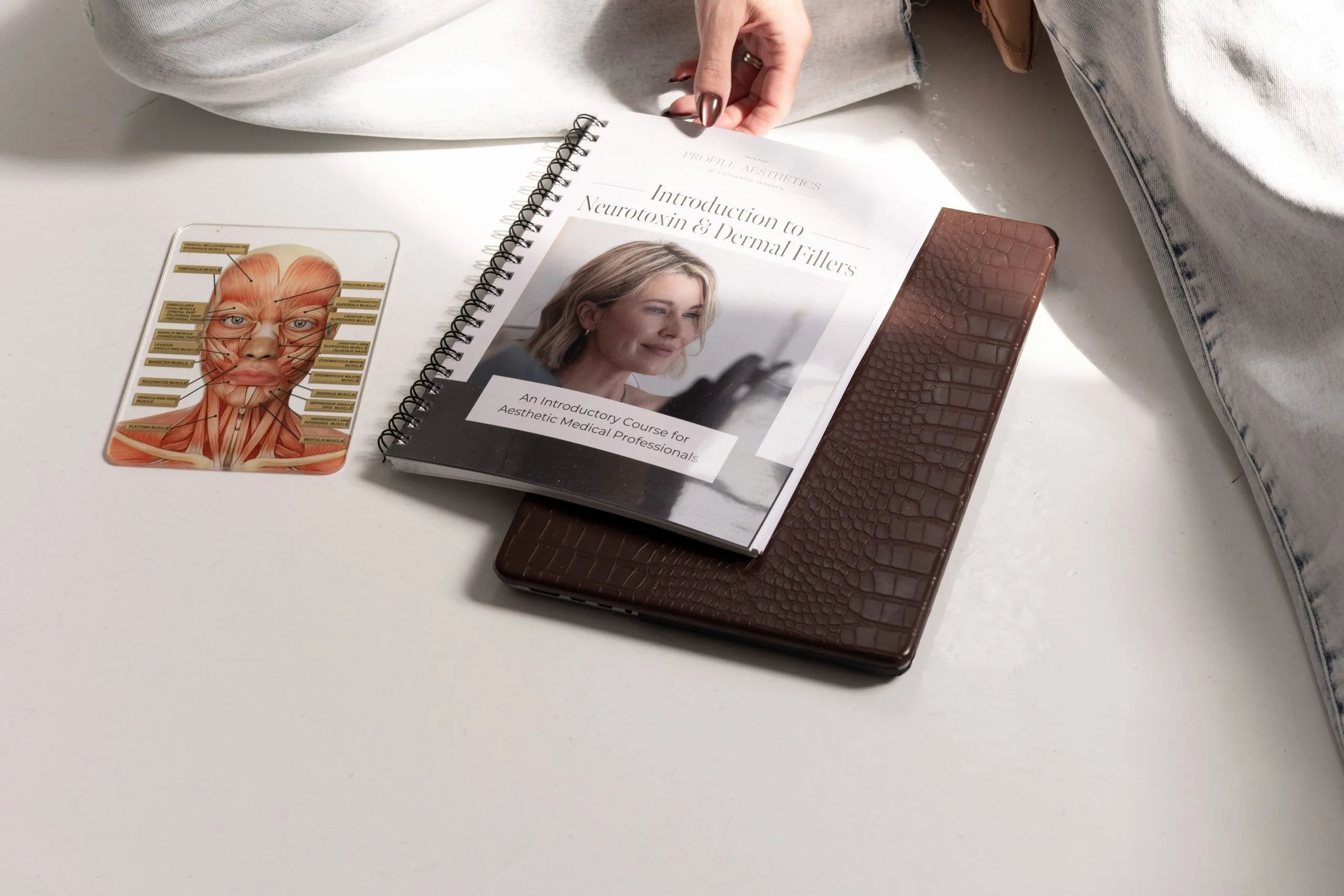 Hand holding a spiral-bound guide titled Introduction to Neurotoxins and Dermal Fillers, displayed with anatomical chart and brown wallet