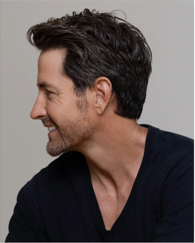 Man in profile view with dark hair and facial hair, wearing navy shirt against neutral background