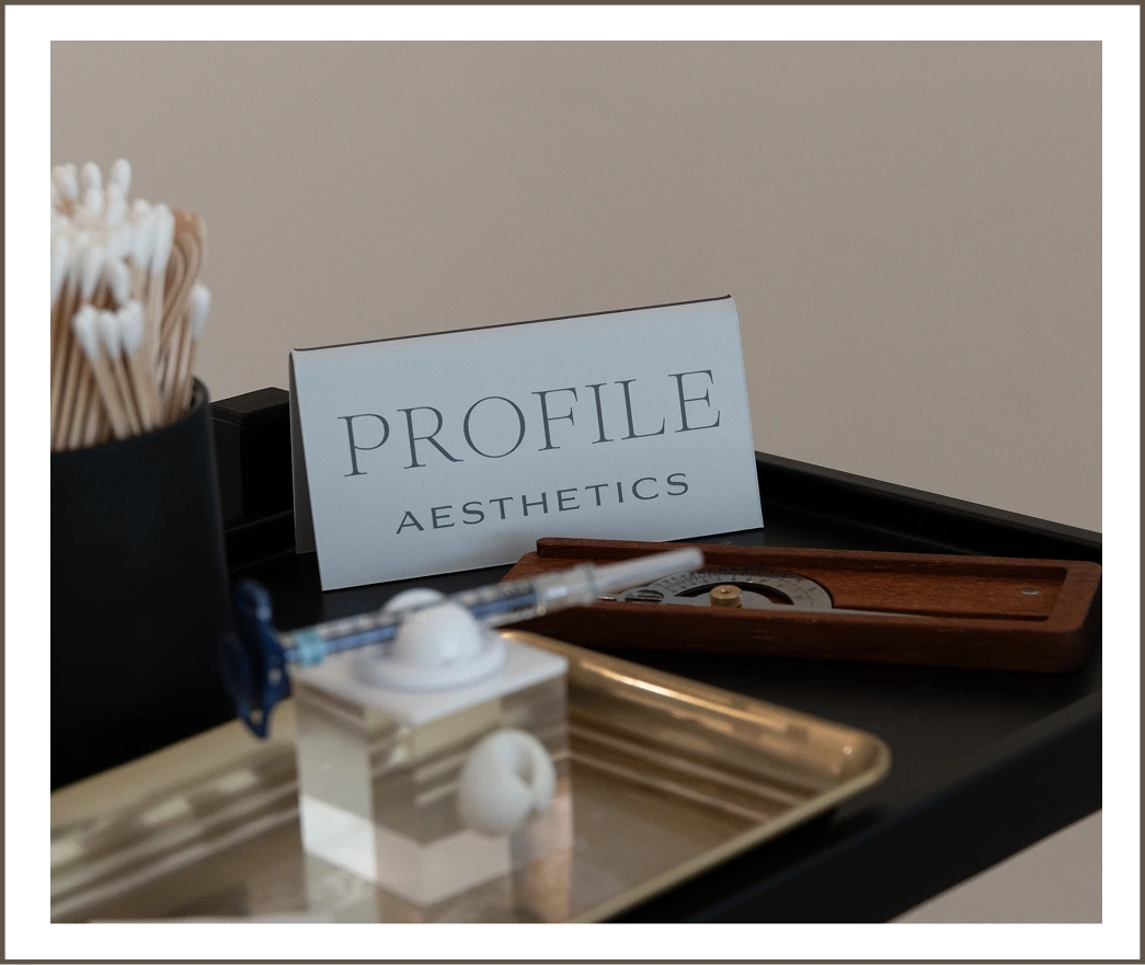 Profile Aesthetics business card displayed on desk with cotton swabs, skincare products, and wooden tray