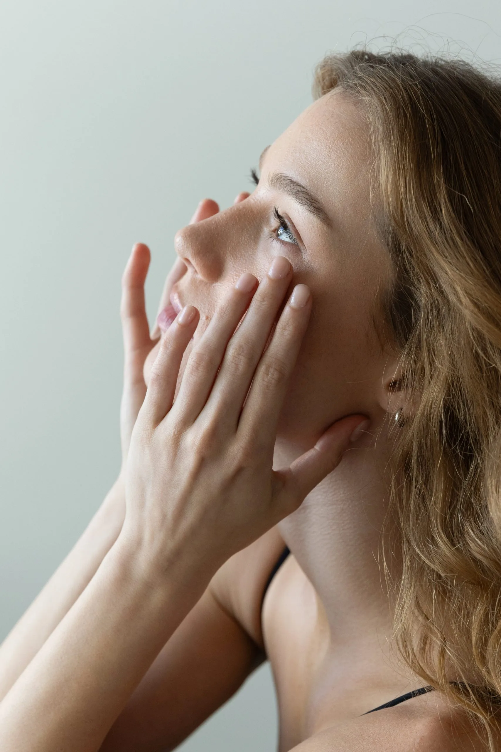 Woman with brown hair touching her face and jawline in profile against a light background