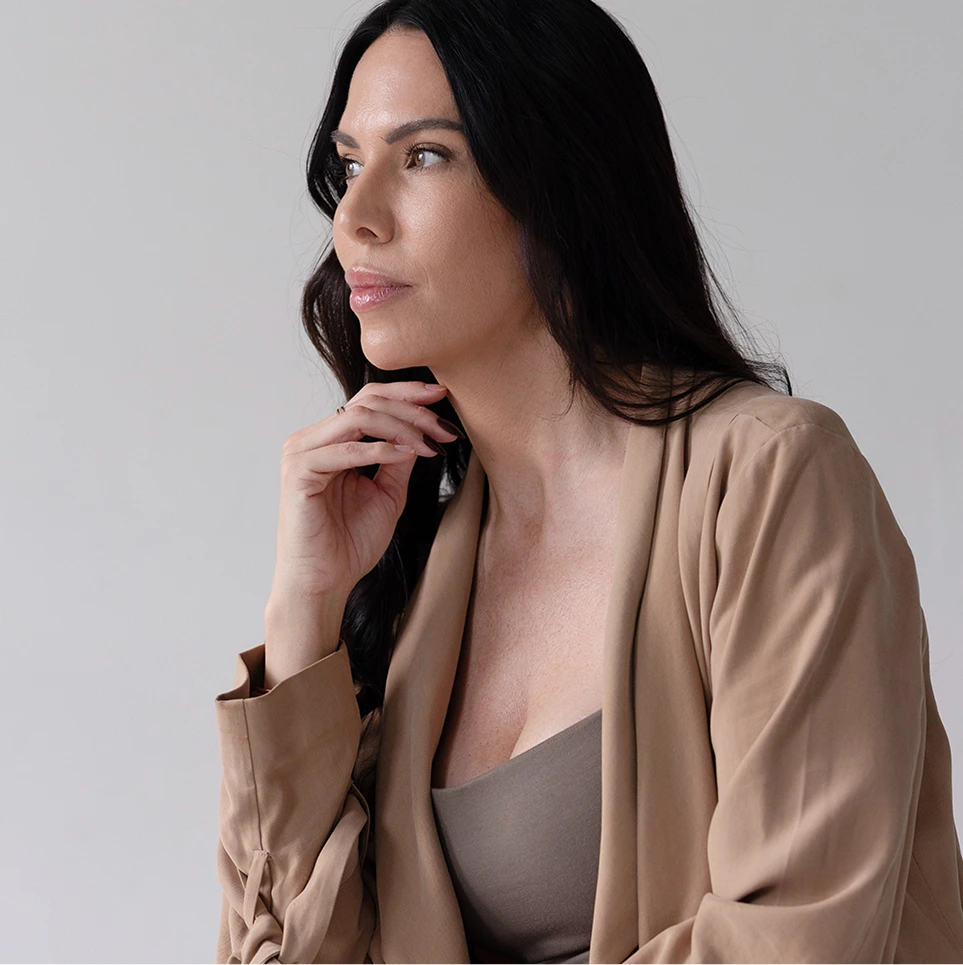 Woman with long dark hair wearing a beige cardigan, hand near chin, looking thoughtfully to the side