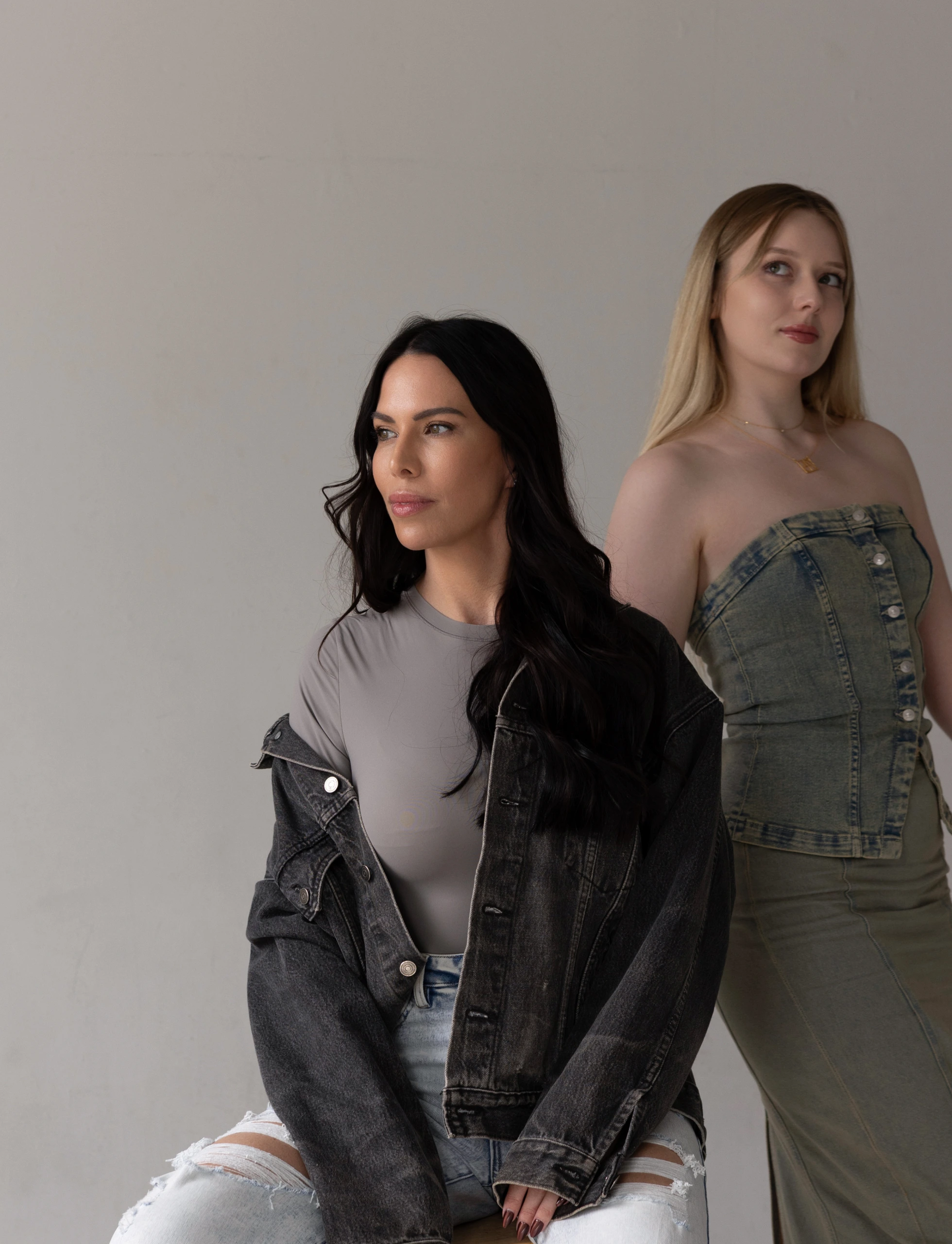 Two women posing against neutral background, one wearing denim jacket with black hair, other in denim top with blonde hair