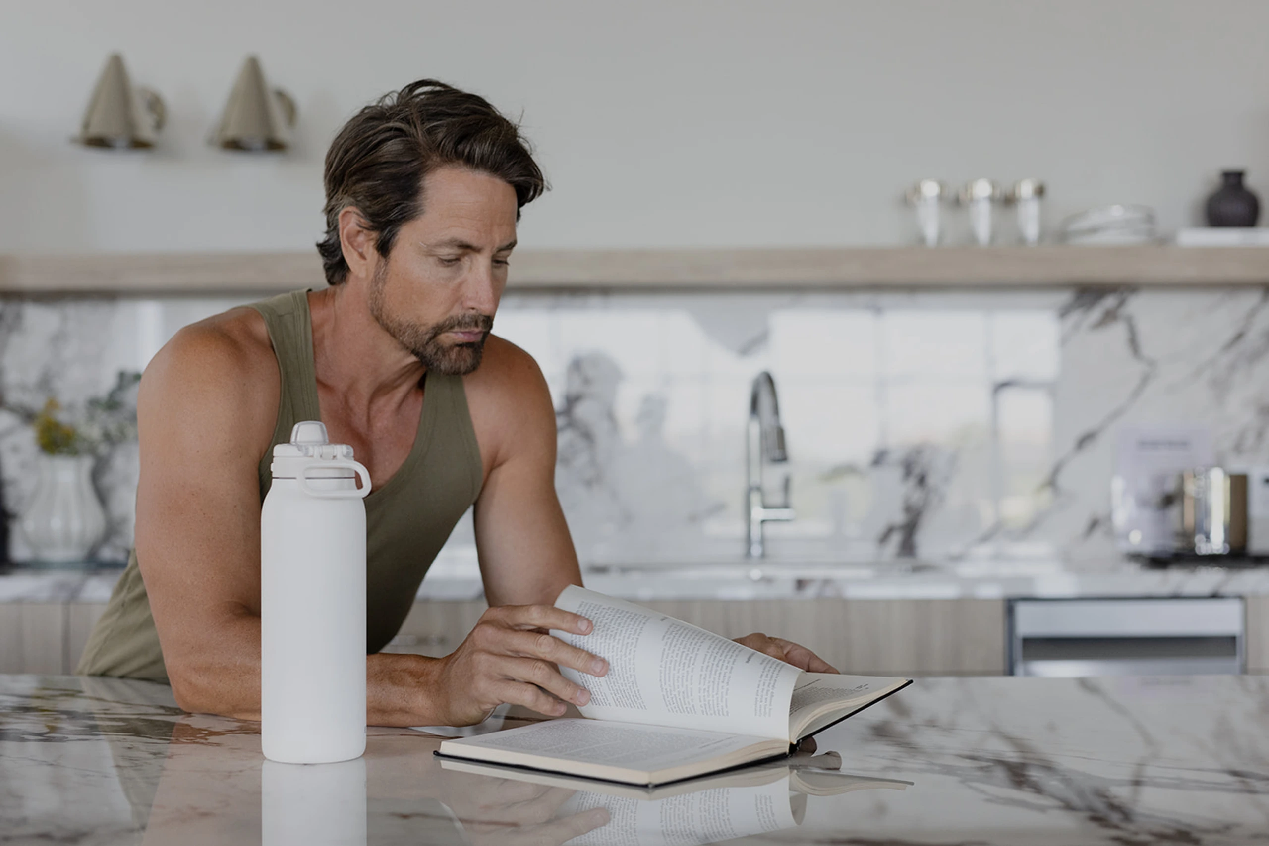 Man in olive tank top reading at kitchen counter next to white recovery product bottle