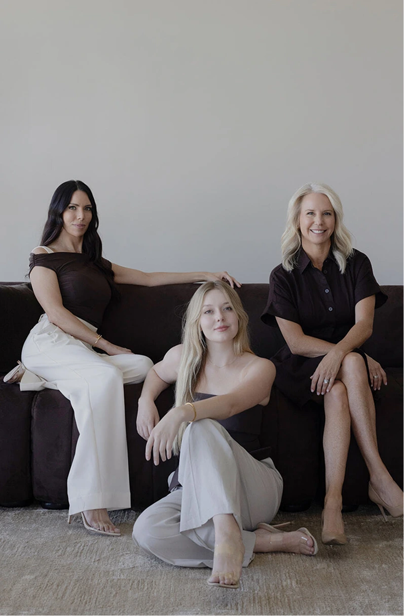 Three women seated on dark couch: woman with long dark hair in black top and white pants on left, woman with blonde hair in neutral tones kneeling in center, woman with blonde hair in black shirt on right
