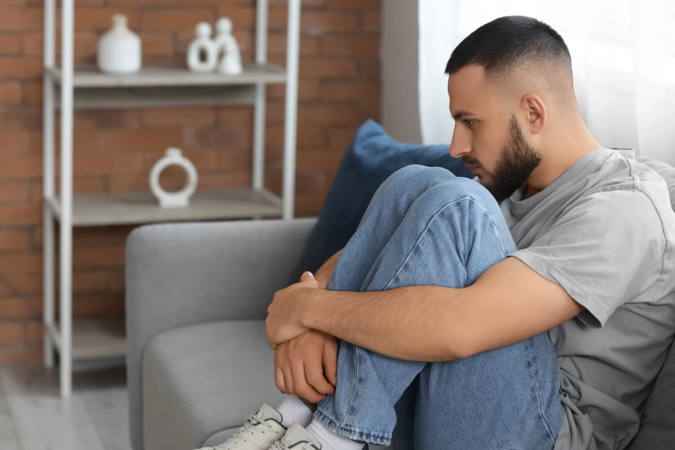 A young man sits alone on a sofa hugging his knees, looking withdrawn and sad