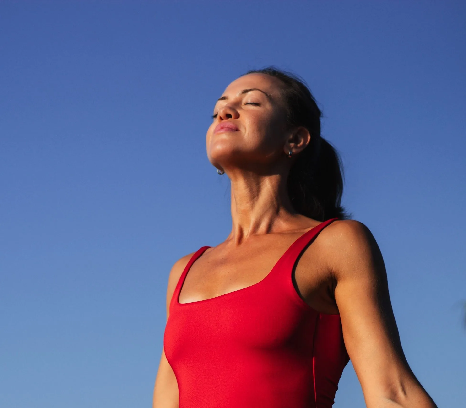 A woman in a red swimsuit tilts her face toward the sun with eyes closed