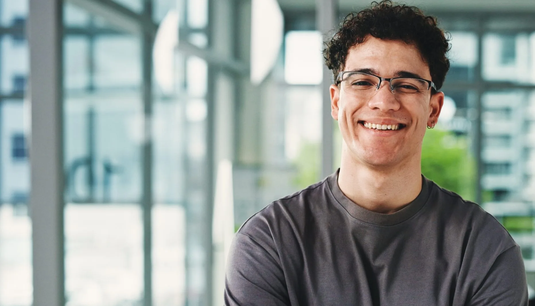 A young man with curly hair and glasses smiles confidently with arms crossed in a bright modern office setting