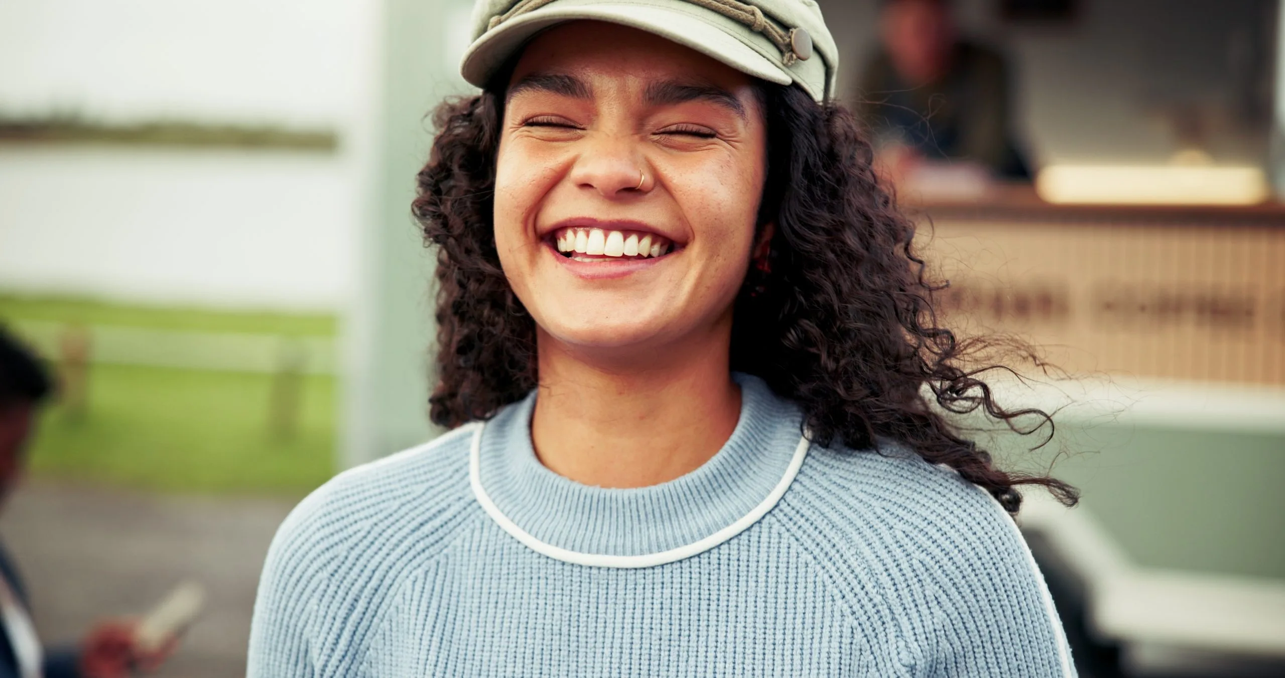 A young woman with curly hair and a cap laughs joyfully outdoors near a food truck