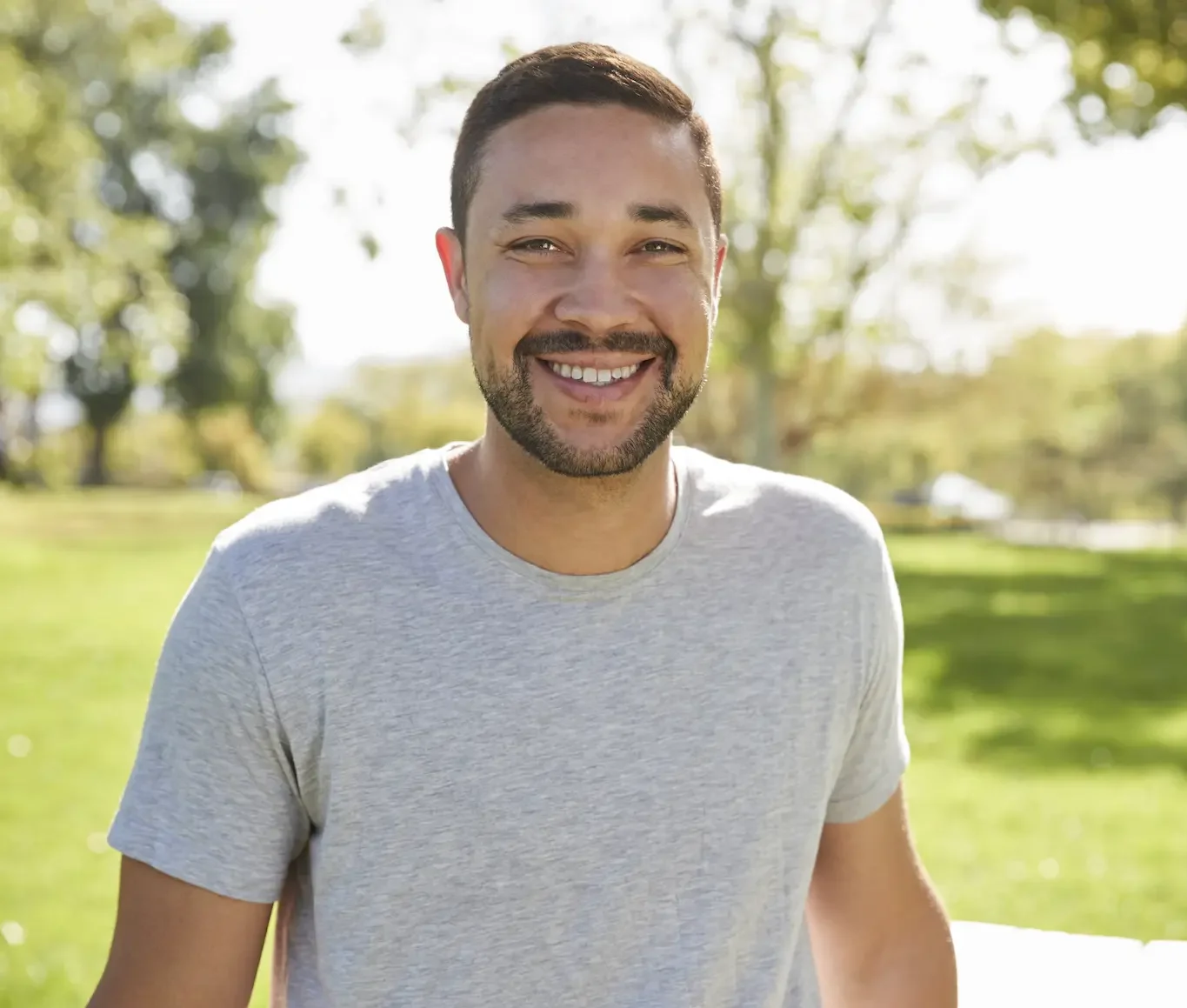 A smiling man in a gray t-shirt poses outdoors in a sunny park setting