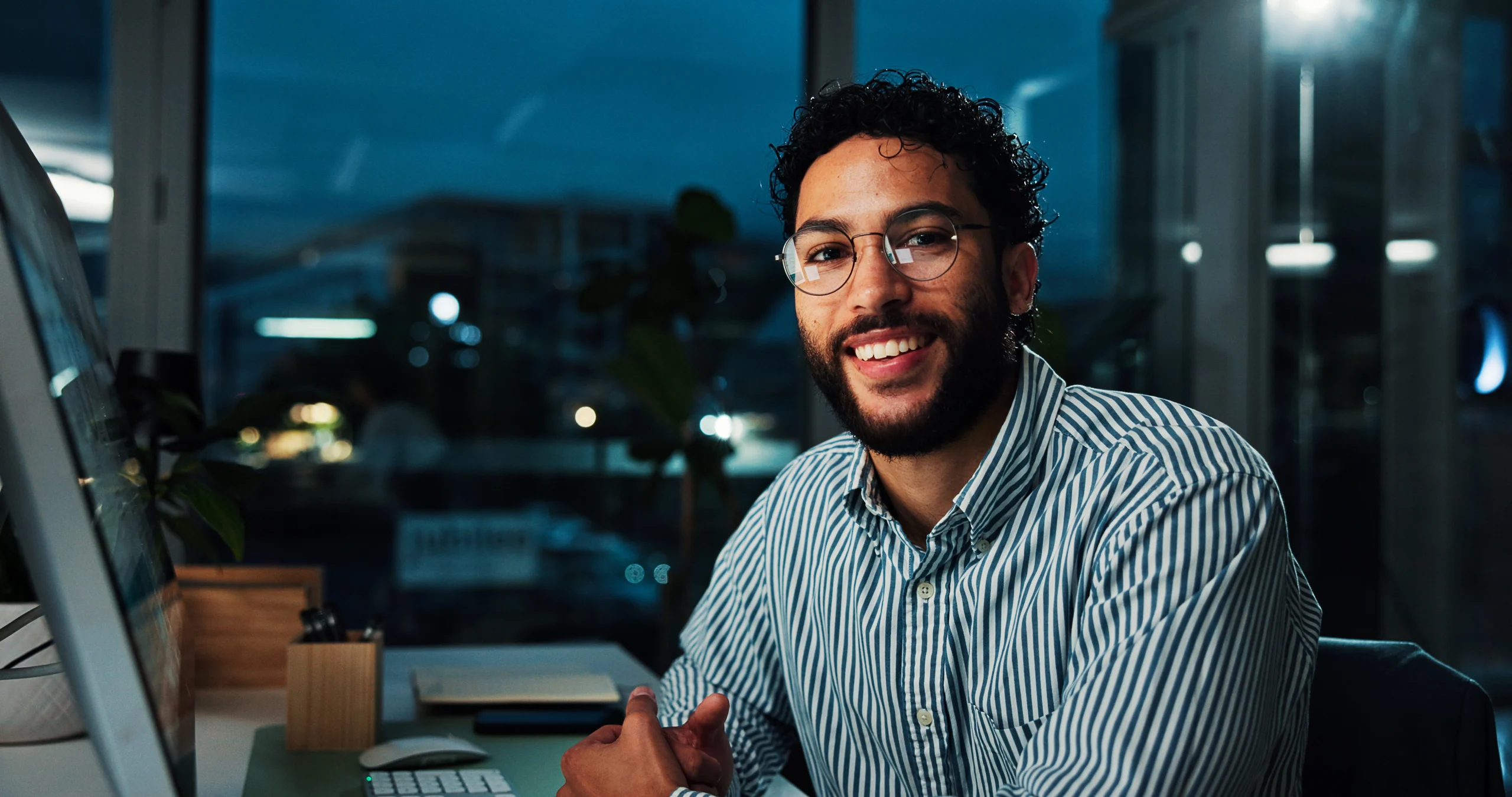 A smiling bearded man wearing glasses and a striped shirt sits at a desk in an office at night with a city vie