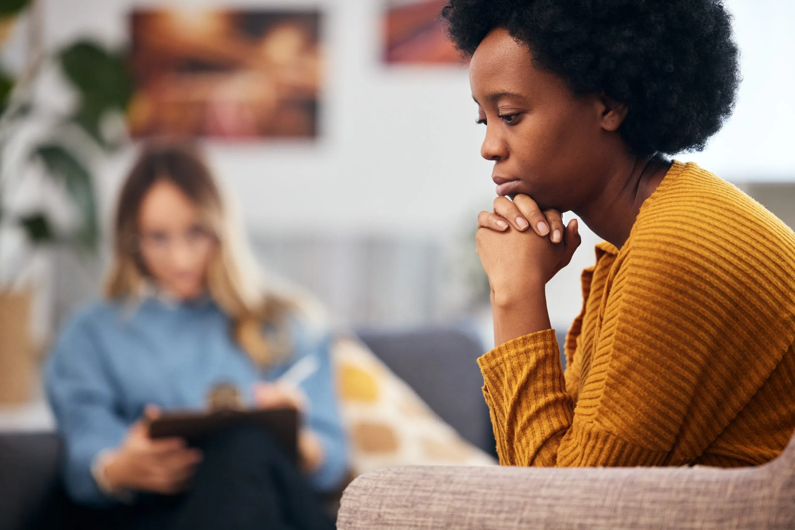 A pensive woman in a mustard sweater sits in a counseling session while a therapist takes notes in the backgro