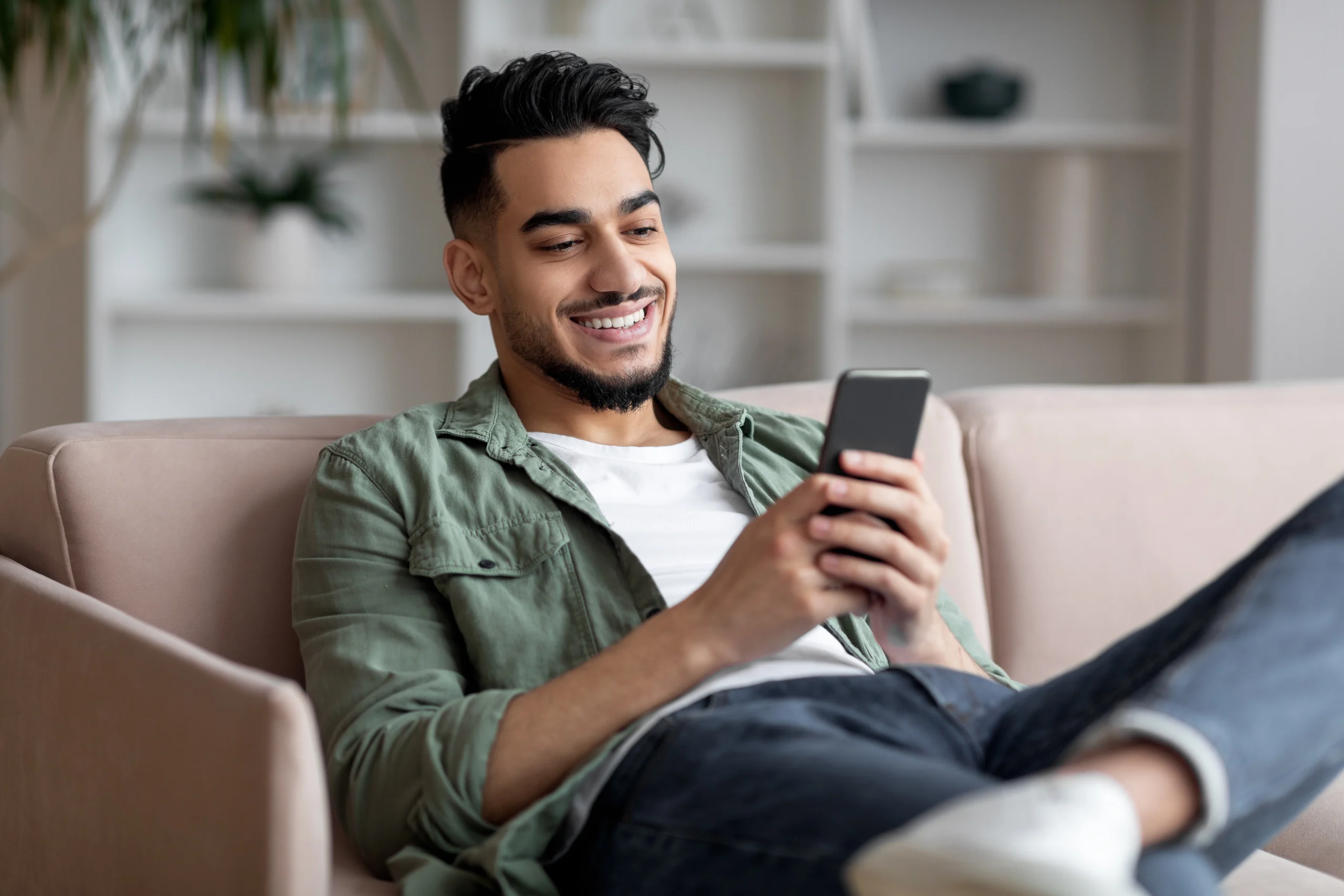 A smiling young man relaxes on a sofa at home while looking at his smartphone