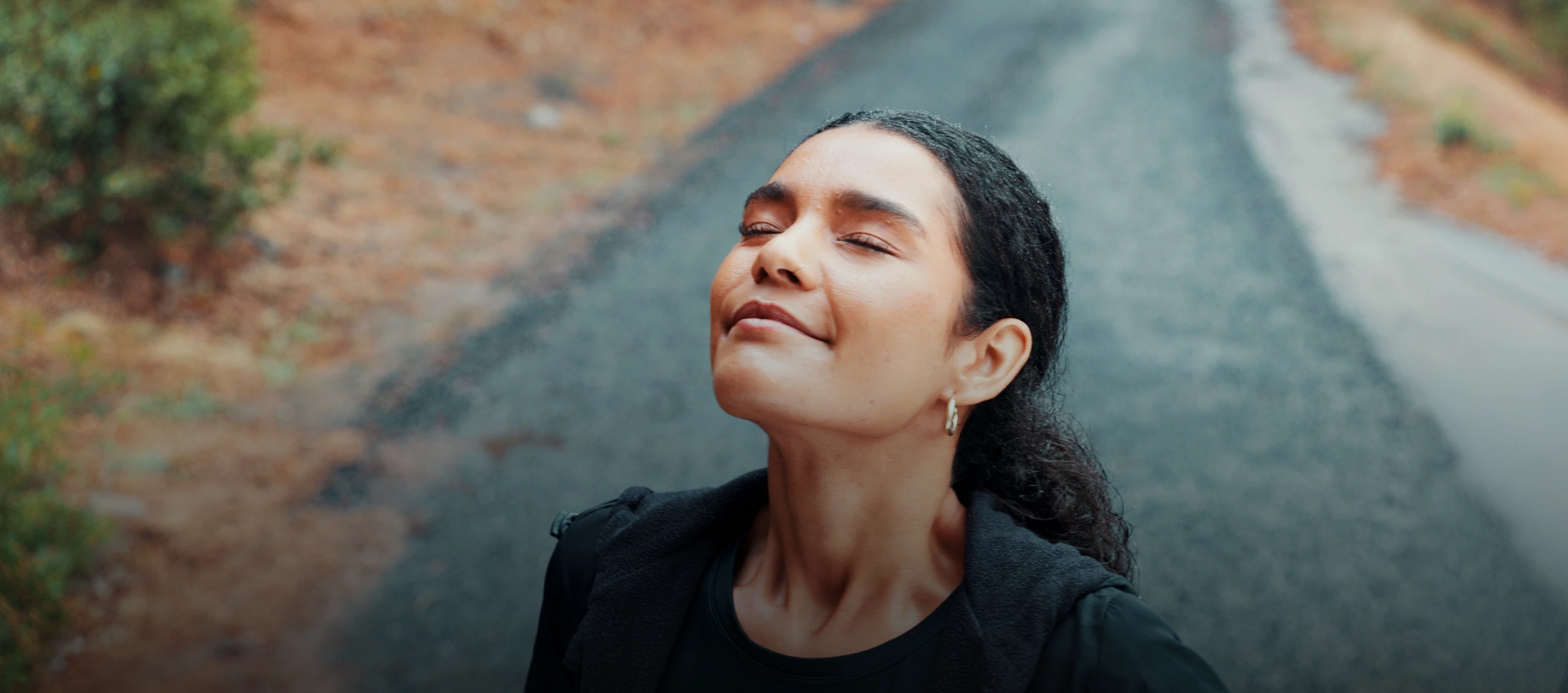 A woman stands on an outdoor path with her eyes closed, tilting her face upward in a moment of calm reflection