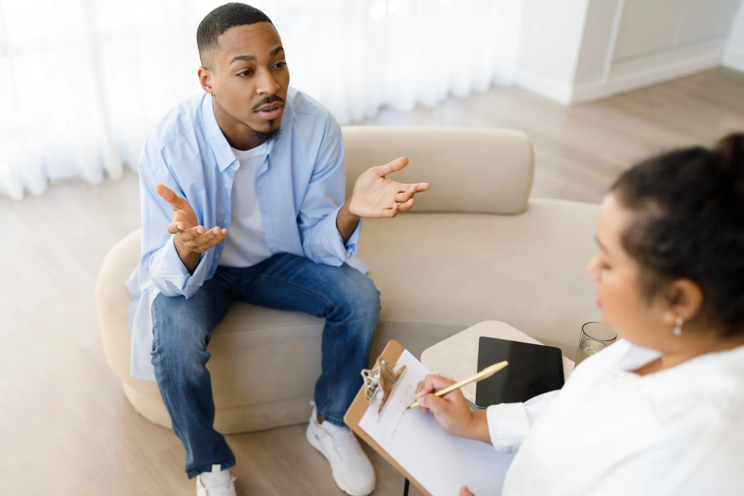 A man gestures while speaking during a therapy session with a clinician taking notes on a clipboard