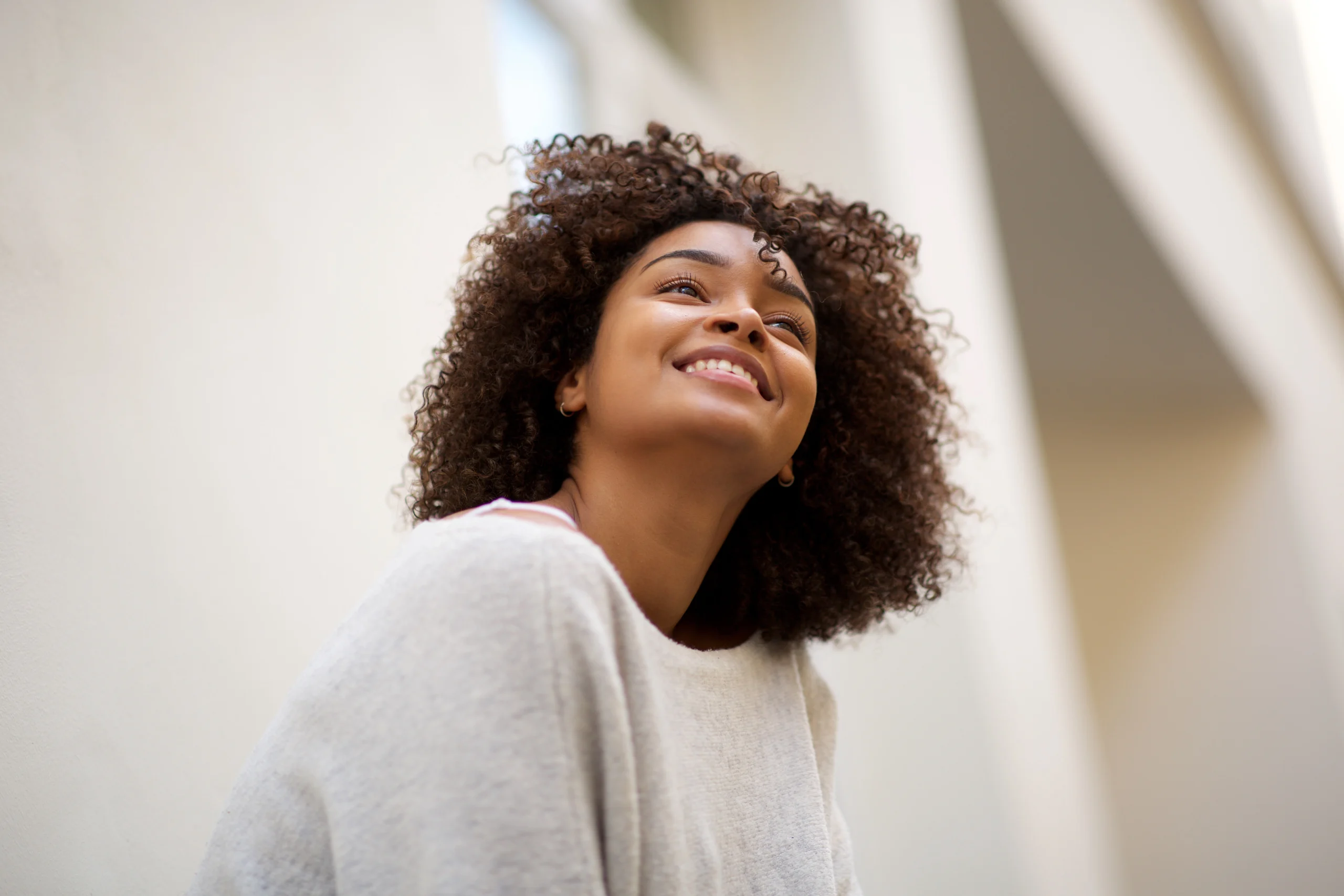A young woman with curly brown hair smiling and looking upward in bright natural light