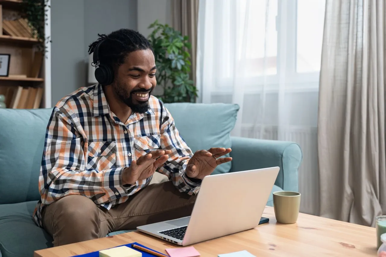A smiling man wearing headphones gestures while having an online therapy video call on his laptop at home