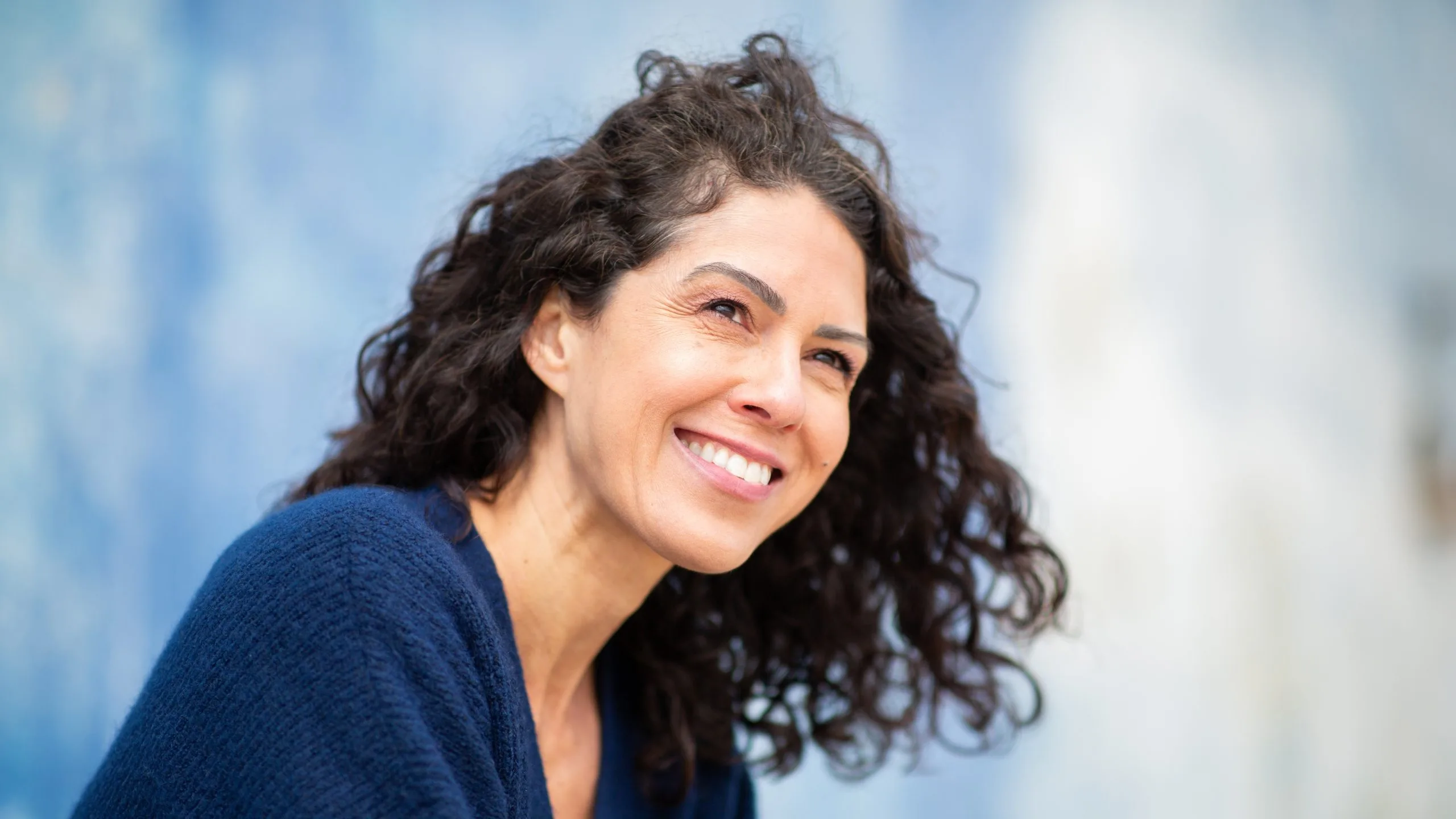 A curly-haired woman in a navy sweater smiles brightly outdoors against a blurred blue background