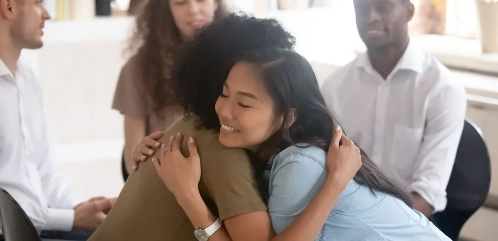 Two women embrace warmly during a group therapy session while other participants look on supportively