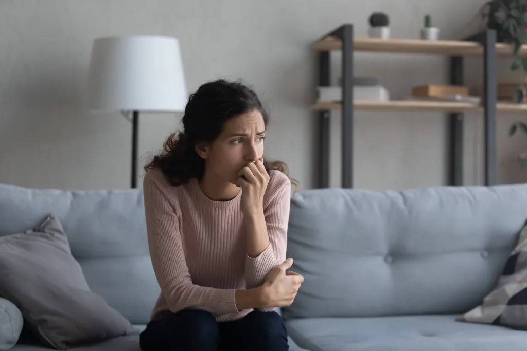 A woman sits anxiously on a sofa at home, biting her nails with a worried expression