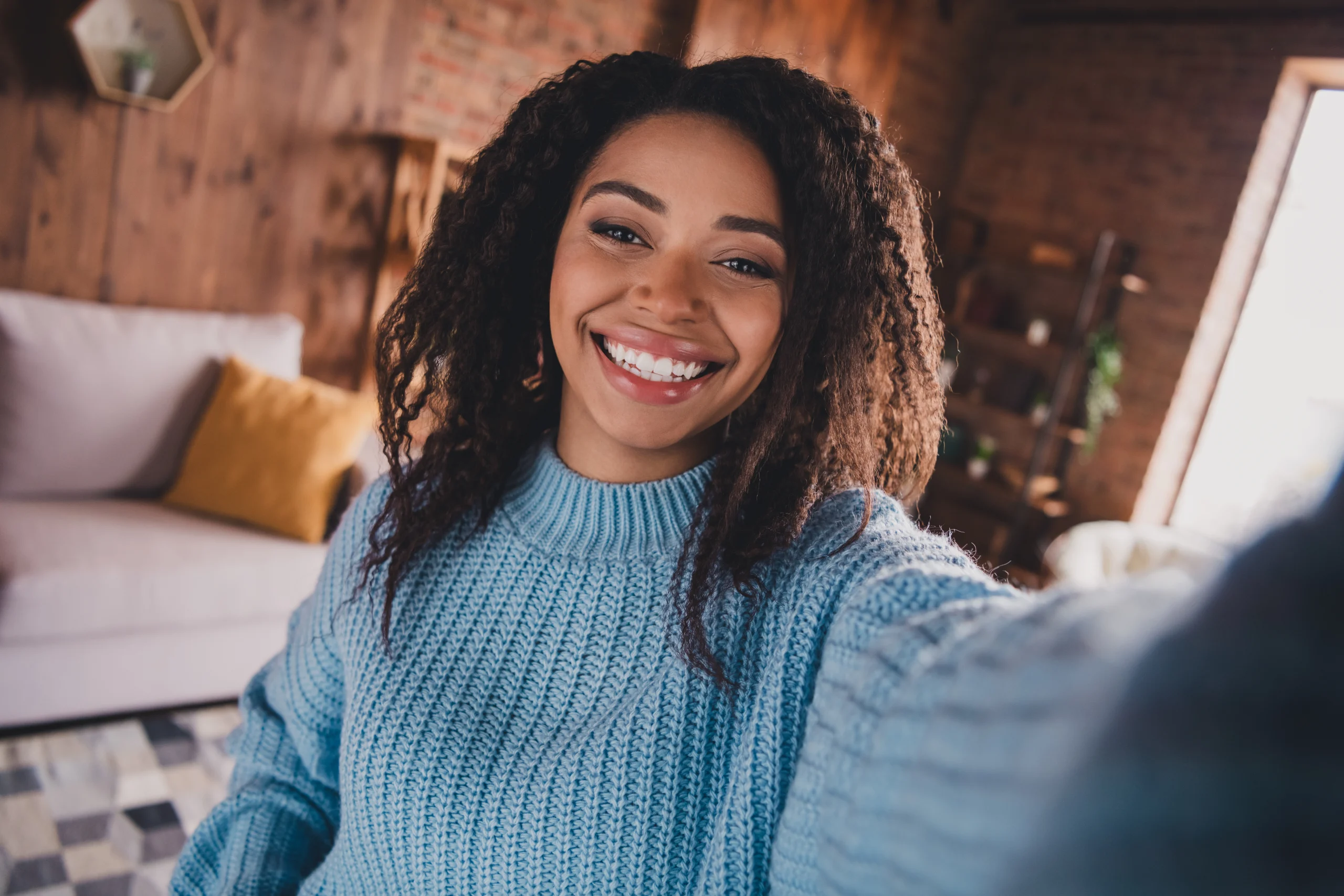 A smiling young woman with curly hair takes a selfie in a cozy living room, wearing a light blue sweater