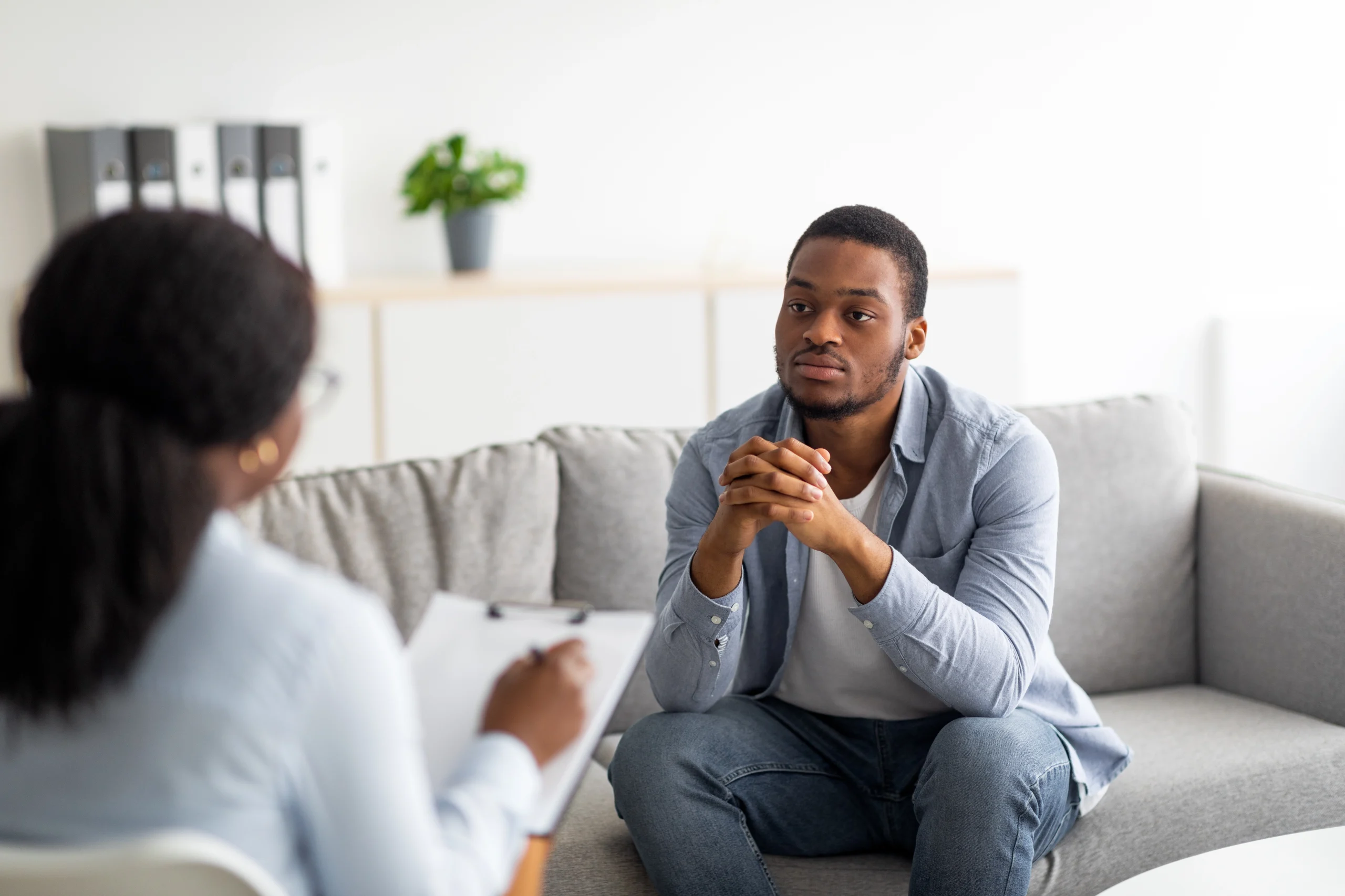 A man sits on a sofa speaking with a clinician who is taking notes on a clipboard during a consultation