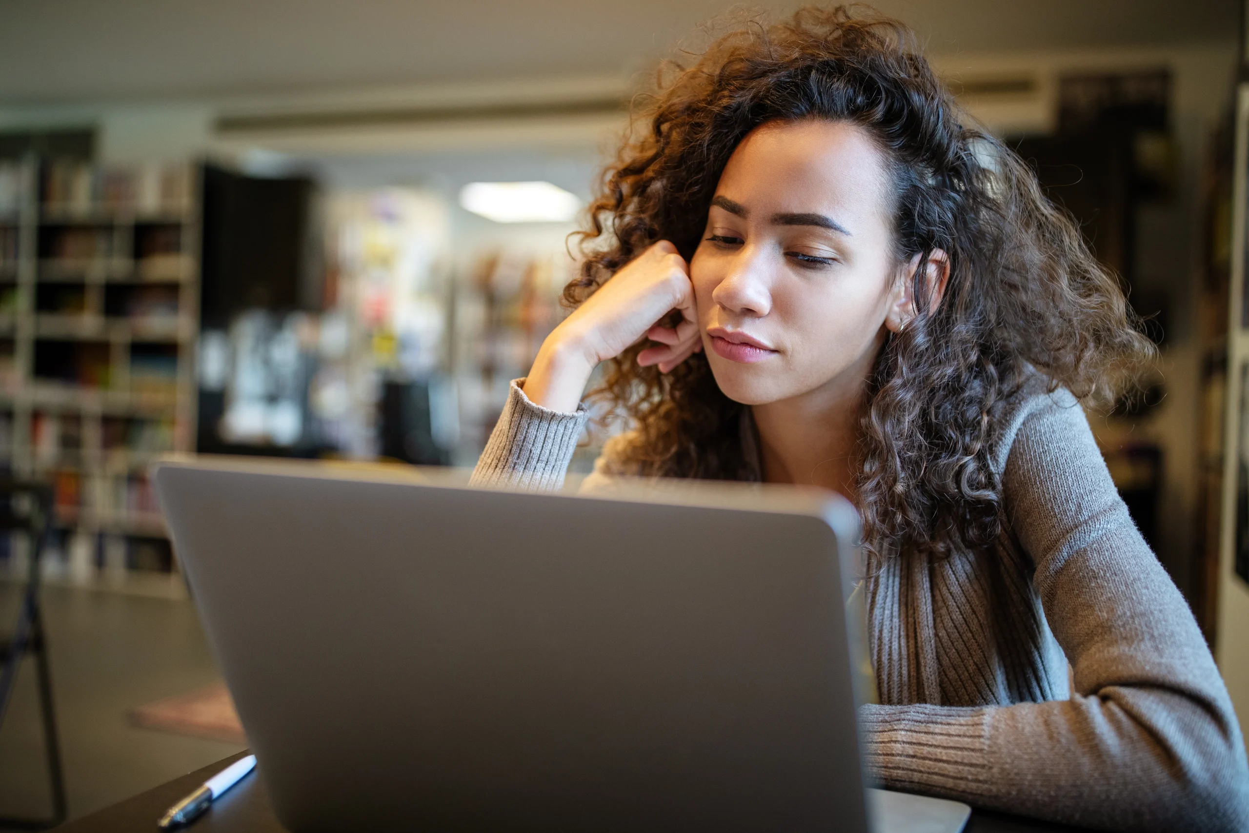 A young woman rests her chin on her hand while looking tiredly at a laptop screen in a library setting