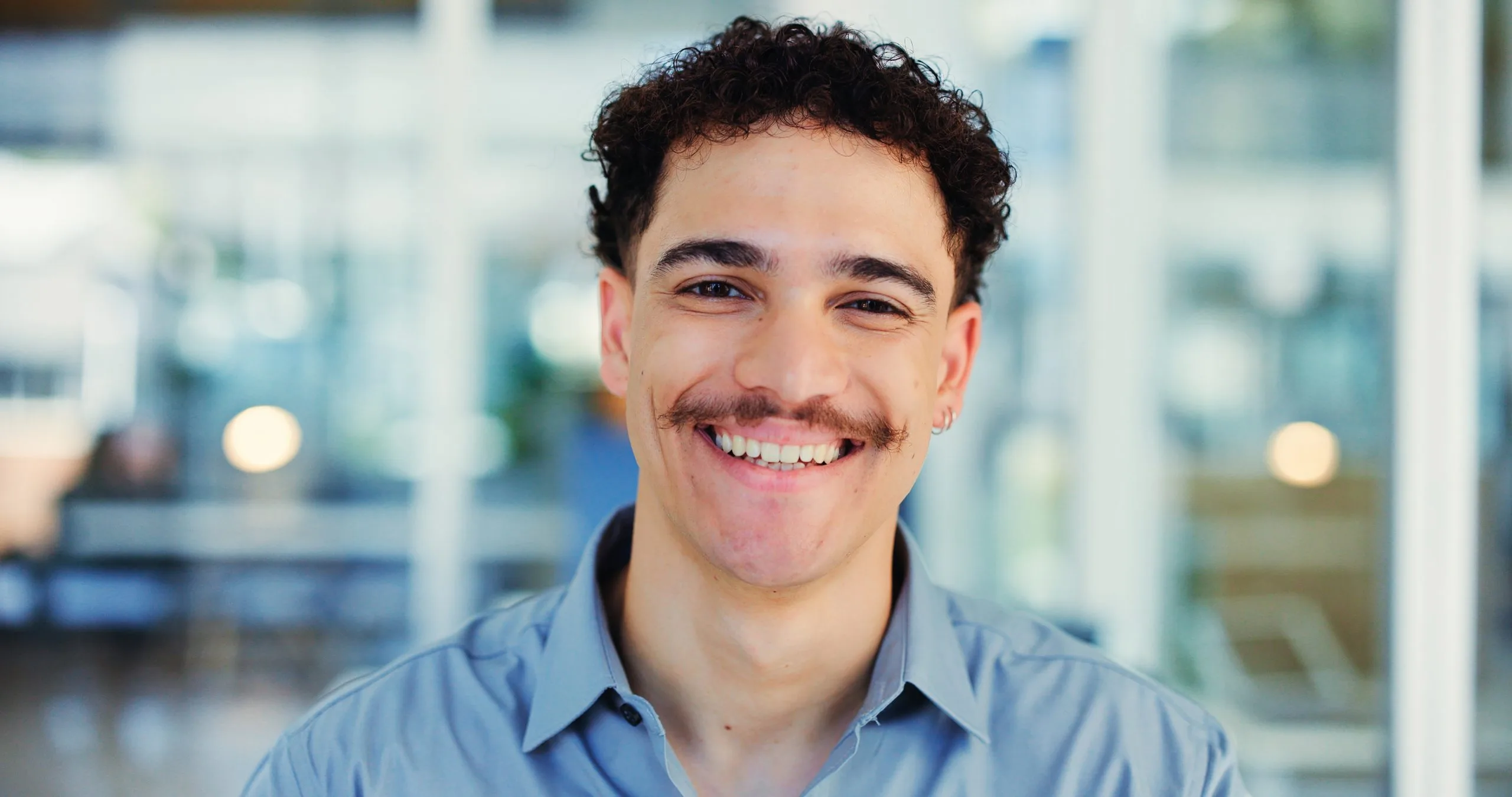 A young man with curly hair and a mustache smiles warmly at the camera in a bright office setting